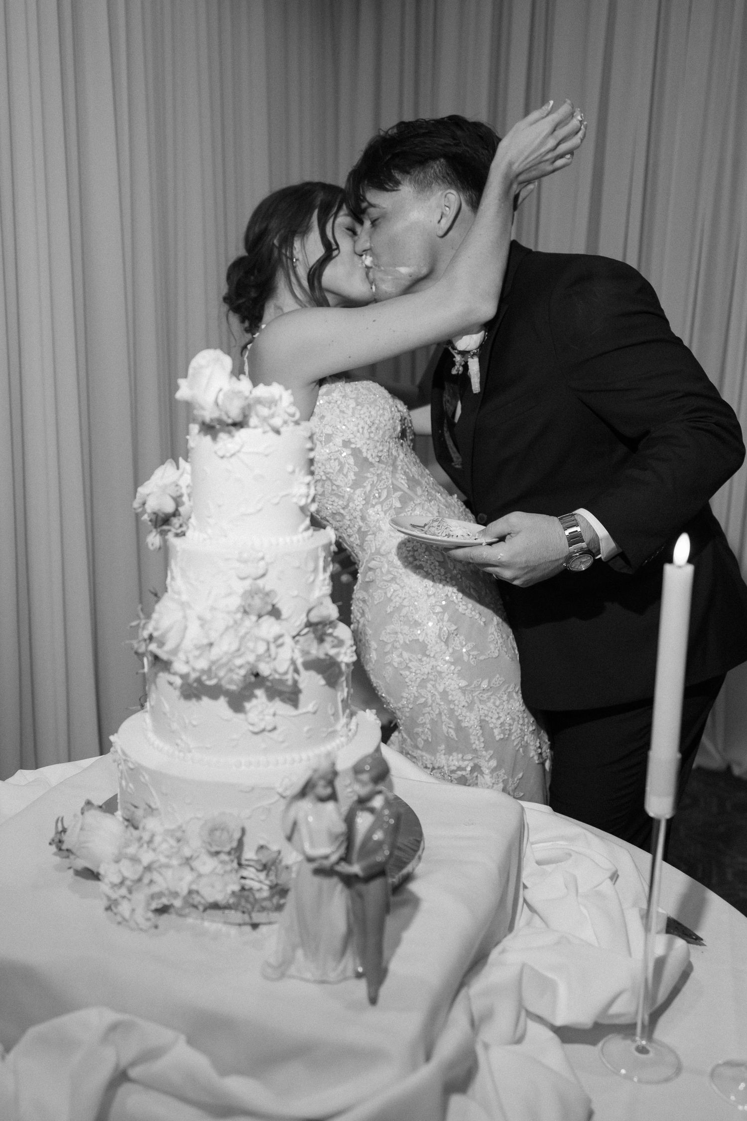 Bride and groom share a kiss beside their elegant multi-tiered wedding cake in a classic black and white photo.