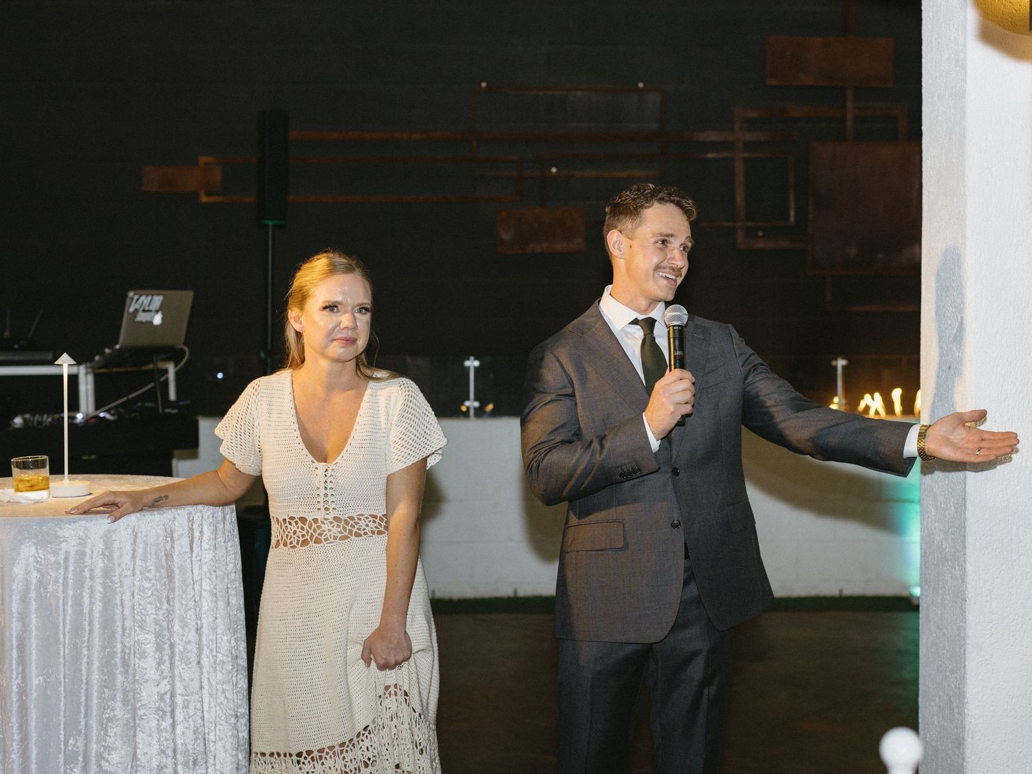A man in a suit speaks into a microphone beside a woman in a white dress at an elegant evening event.