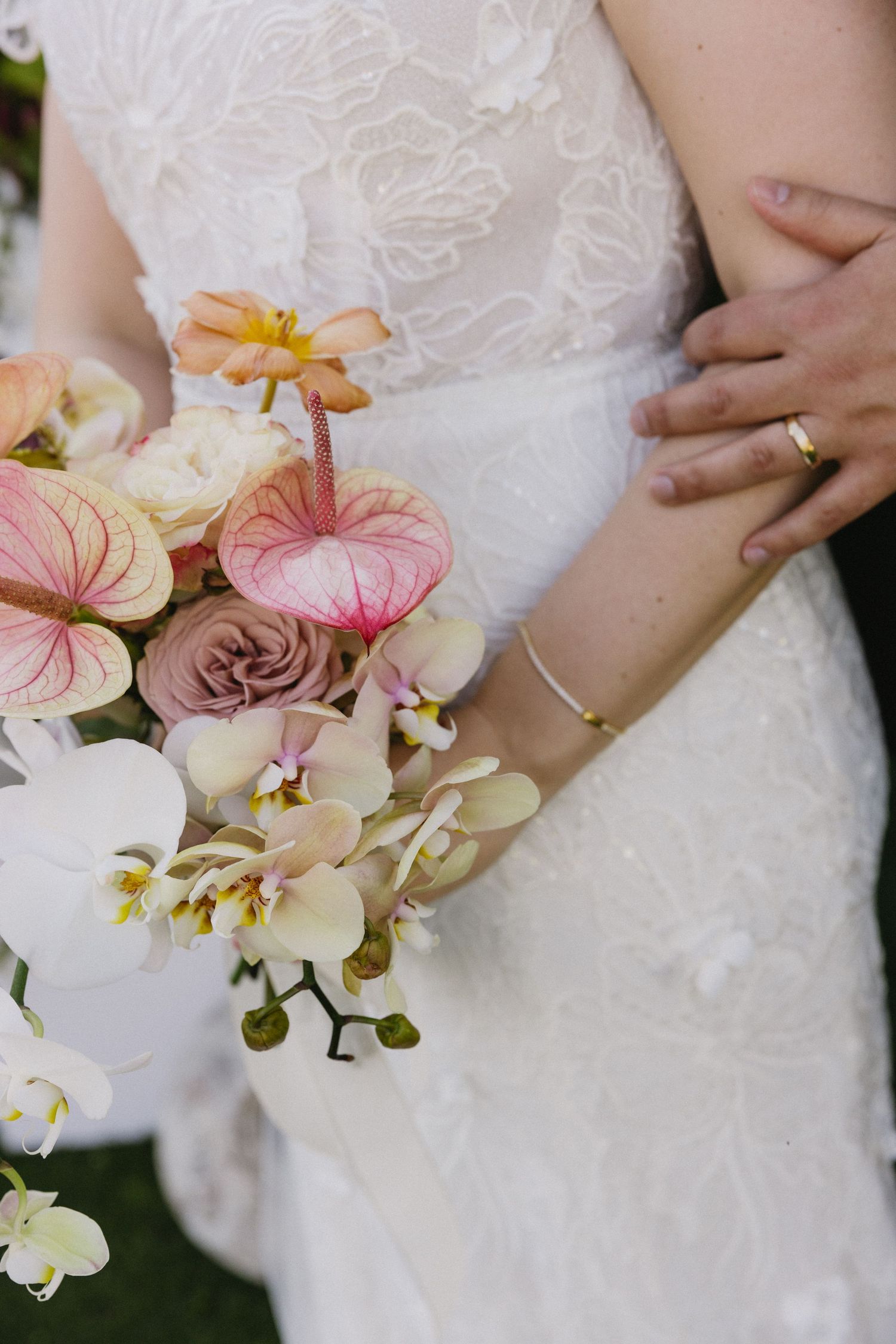 Bride in lace wedding dress holding bouquet of pink anthuriums, roses, and white orchids while groom embraces her waist.