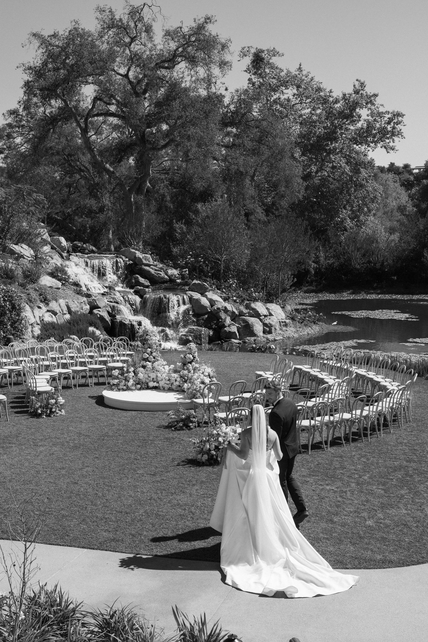 Black and white photo of bride and groom walking toward outdoor wedding ceremony with waterfall backdrop.