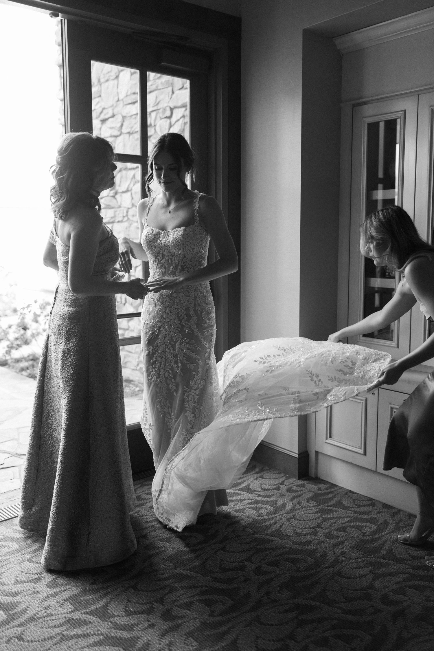 Black and white photo of a bride in lace gown being helped by bridesmaids near a window on wedding day.