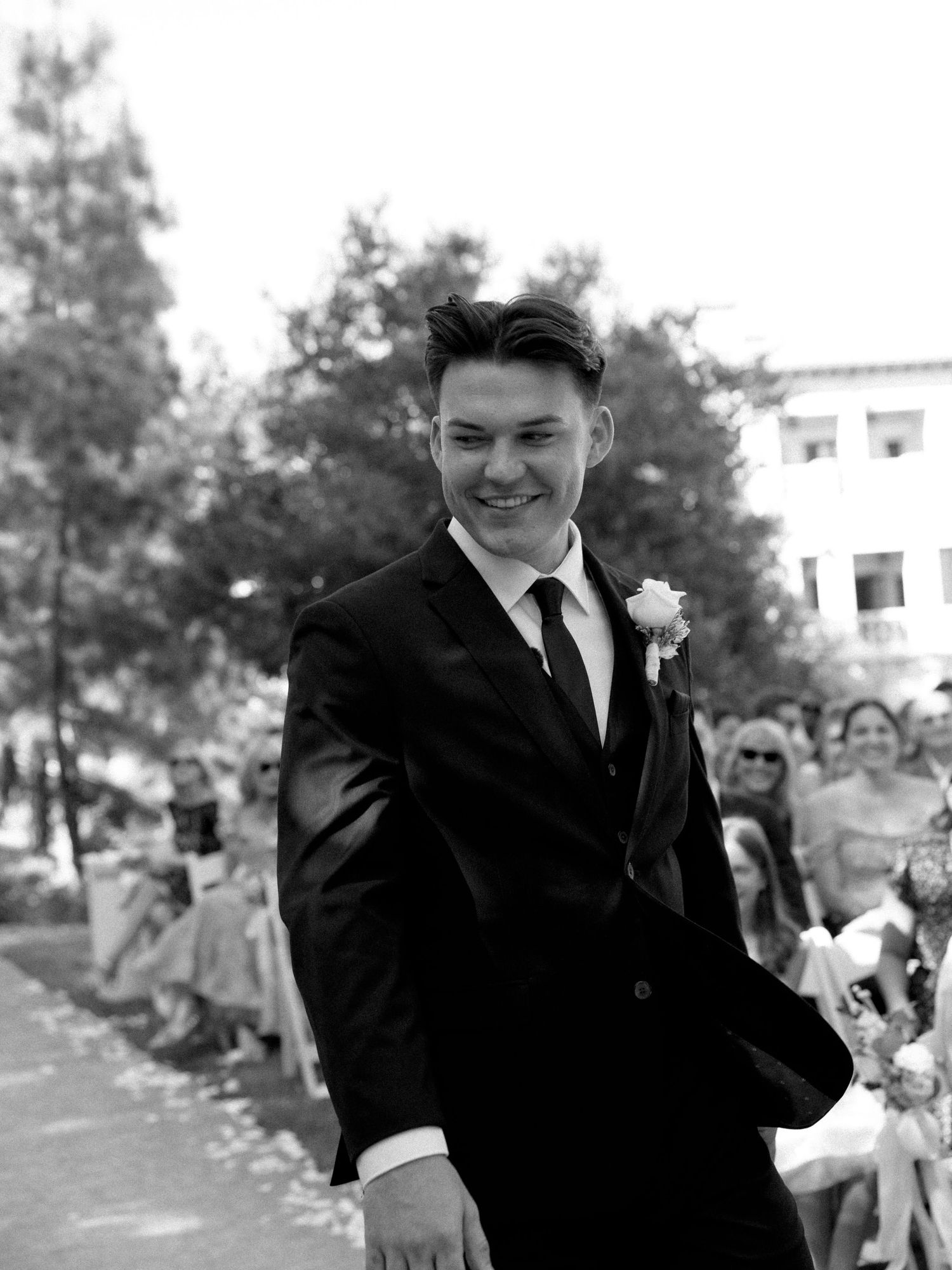 Young man in black suit with boutonniere smiling at outdoor wedding ceremony, black and white photo.
