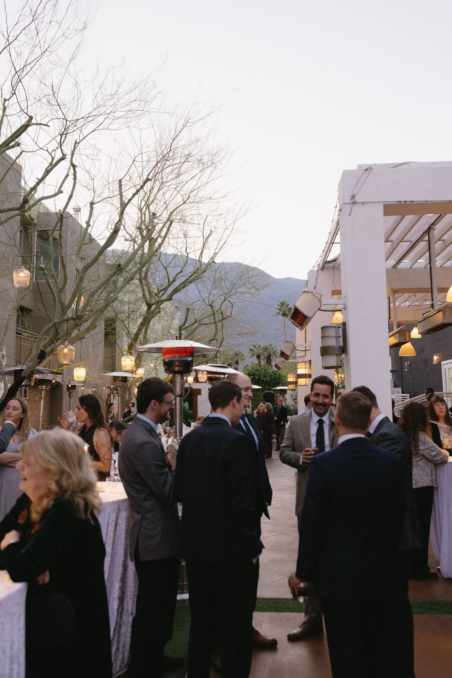 Elegant outdoor wedding cocktail hour with guests mingling along a courtyard pathway, mountain views and patio heaters at dusk.