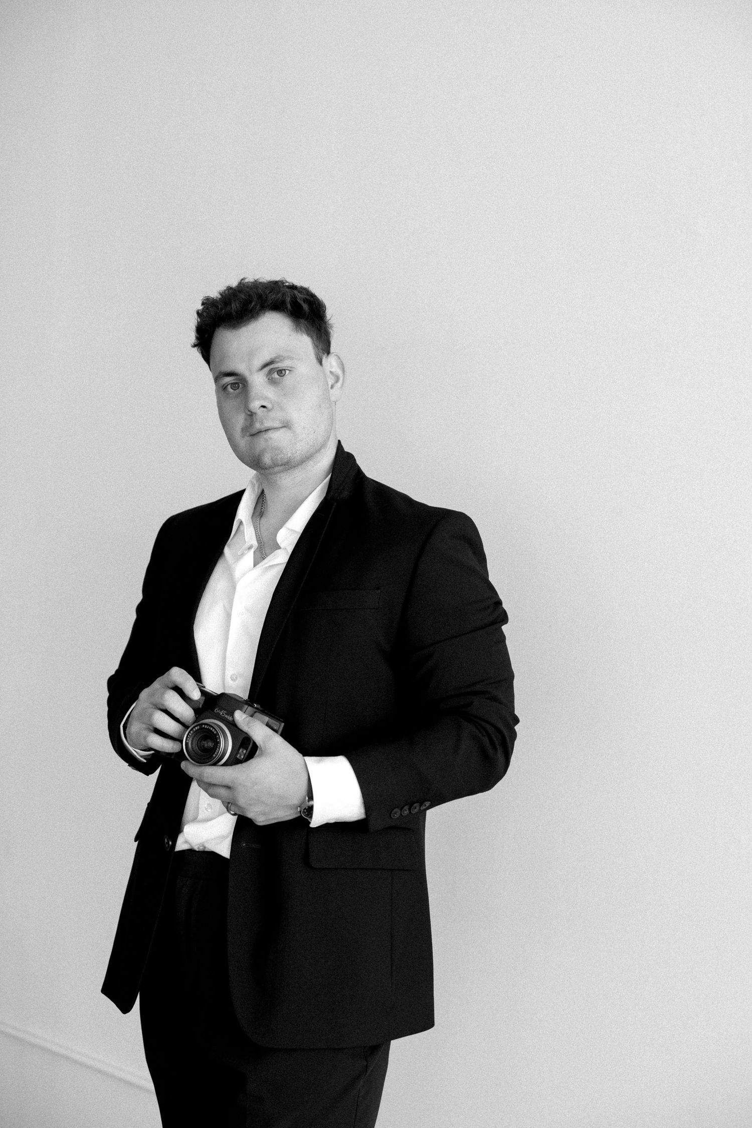 Black and white portrait of a man in a suit holding a camera, standing against a light background.