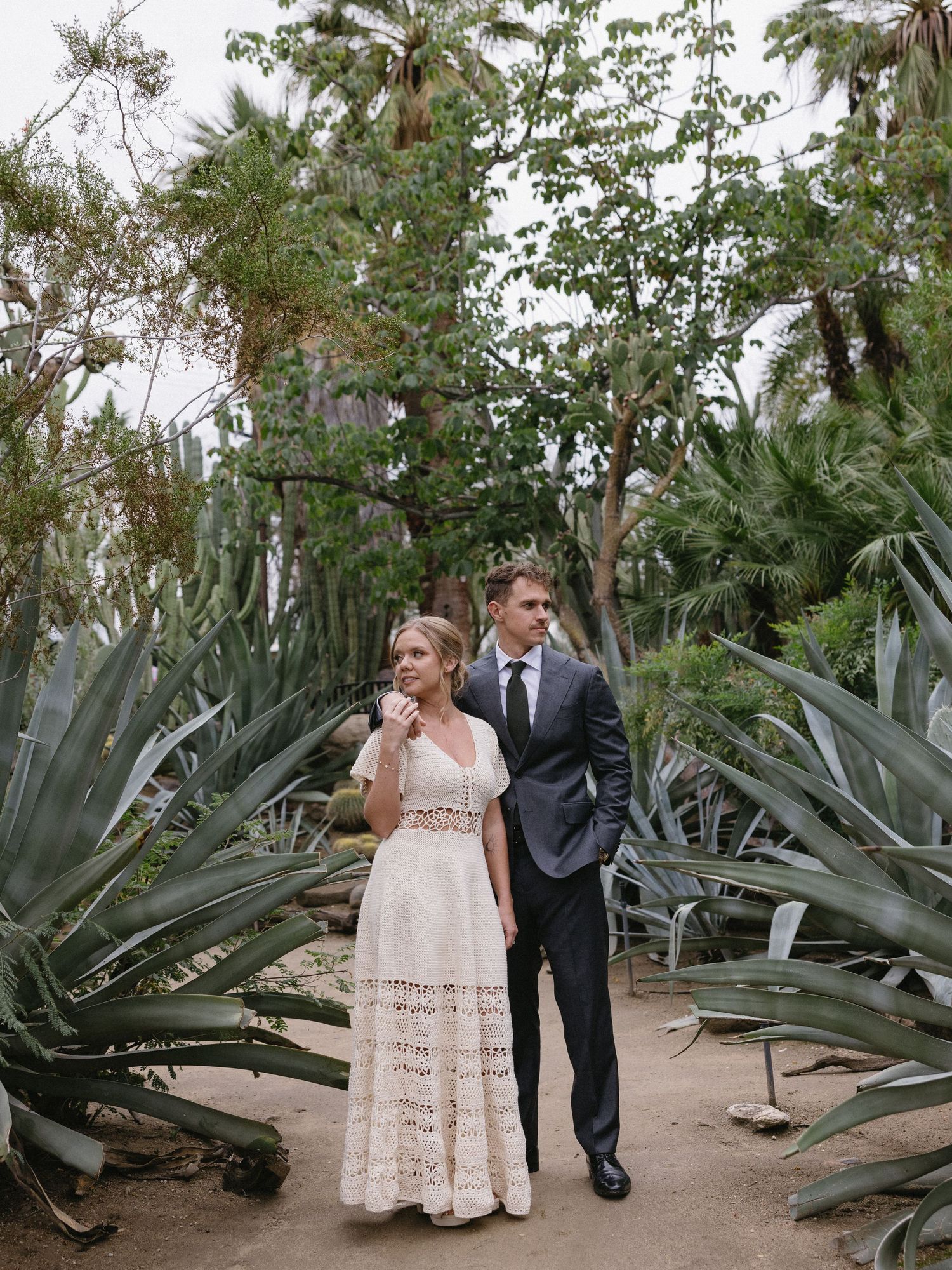 Bride in lace gown and groom in dark suit walk among towering cacti and palm trees in a lush garden setting.
