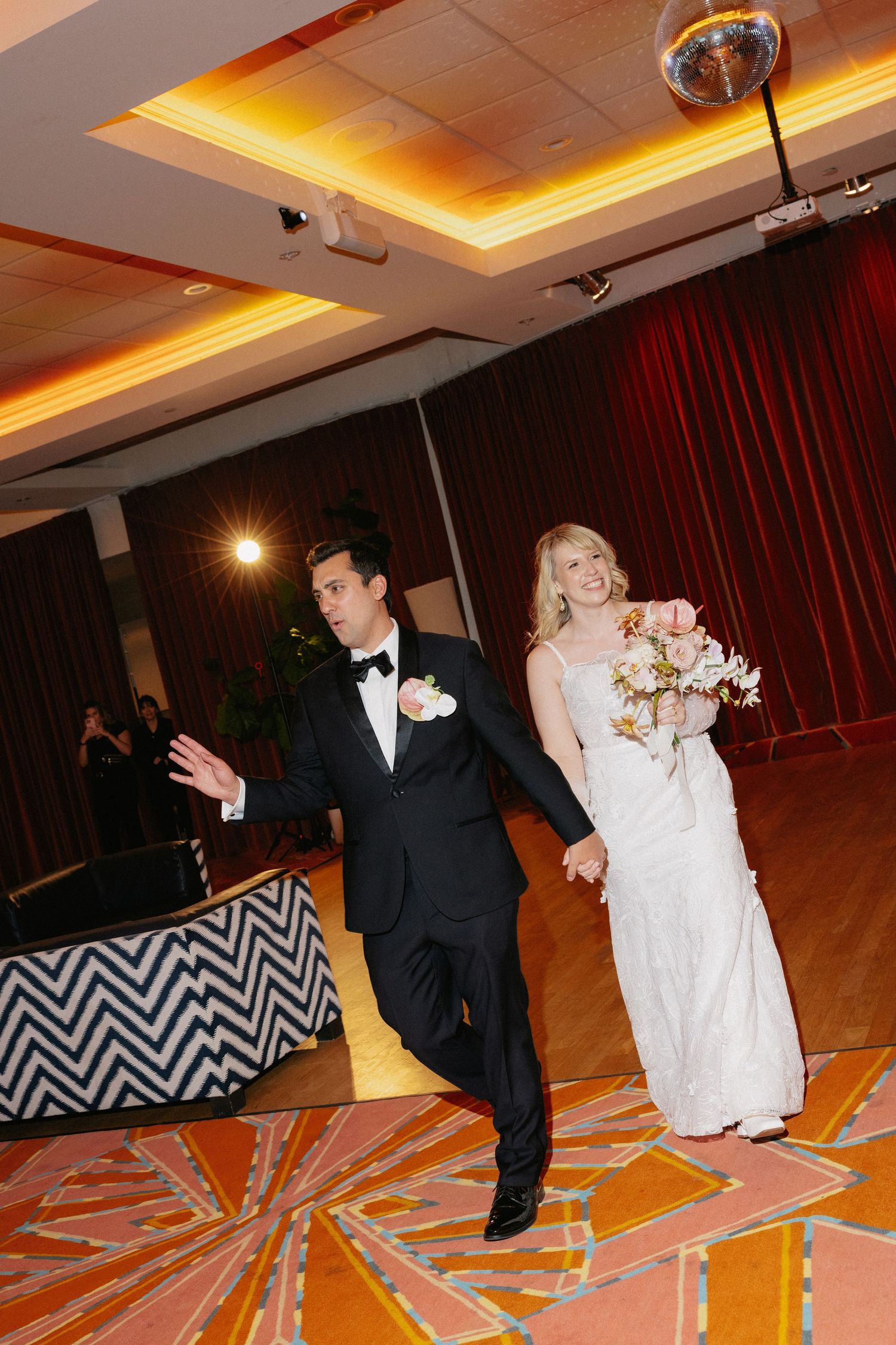 Bride and groom make grand entrance at wedding reception, she holds bouquet in white gown, he gestures in black tuxedo.