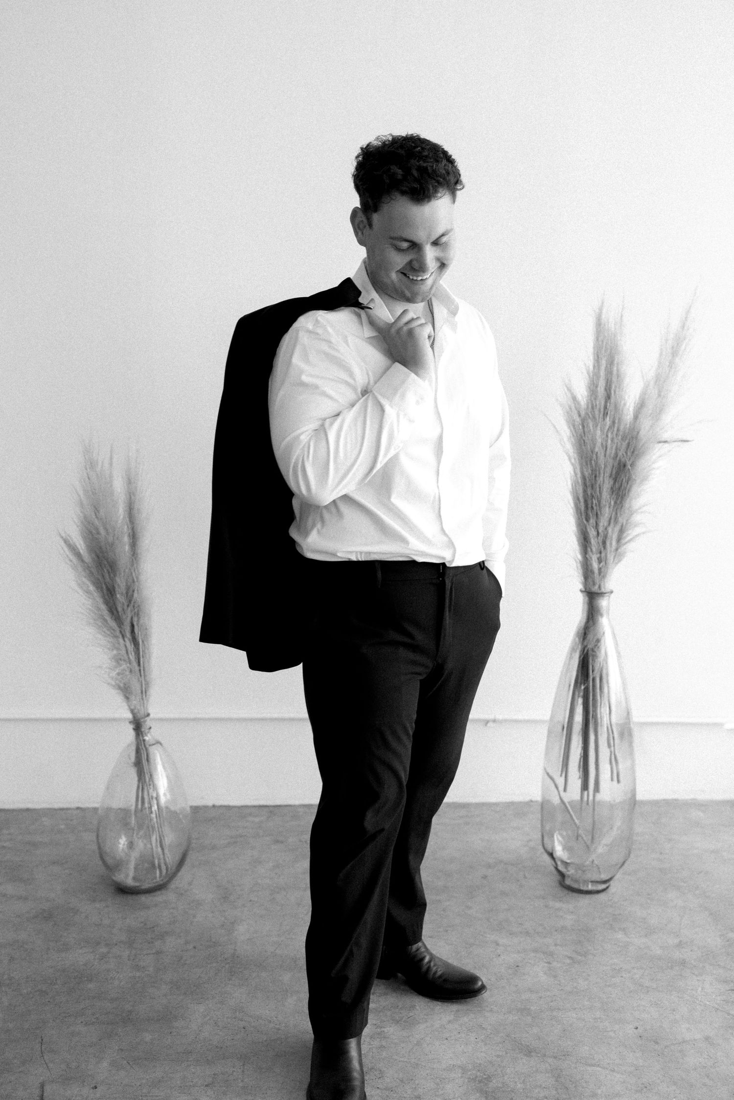 A man in a white dress shirt and black trousers holds a jacket over his shoulder beside tall vases with dried pampas grass.