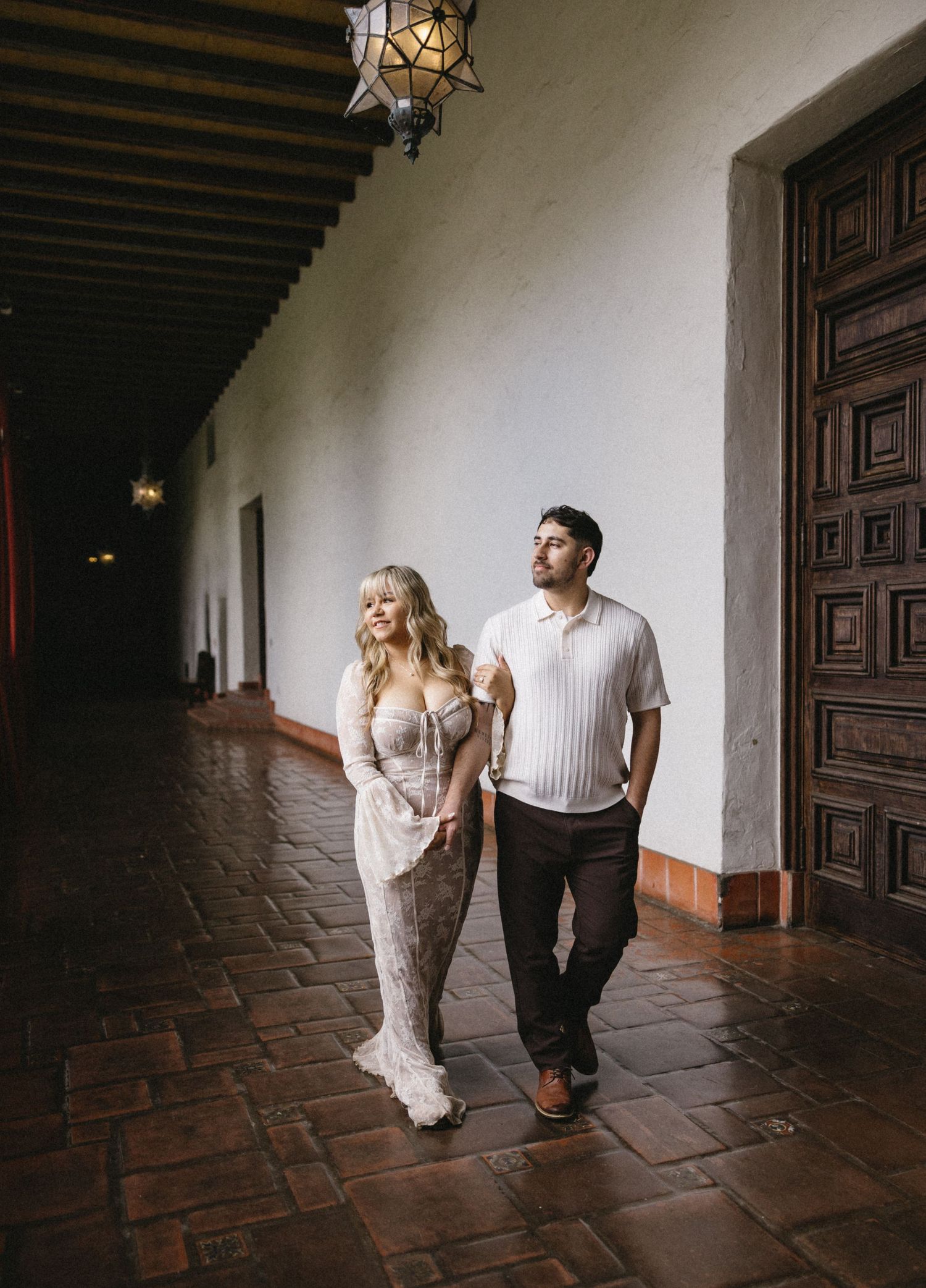 A couple strolls down a dimly lit hallway with terracotta floors and Spanish colonial architecture.