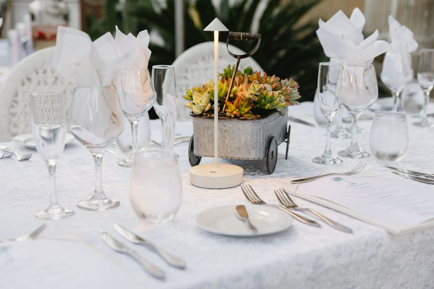 Elegant white wedding reception table setting with floral centerpiece, crystal glasses, and silverware on a white tablecloth.