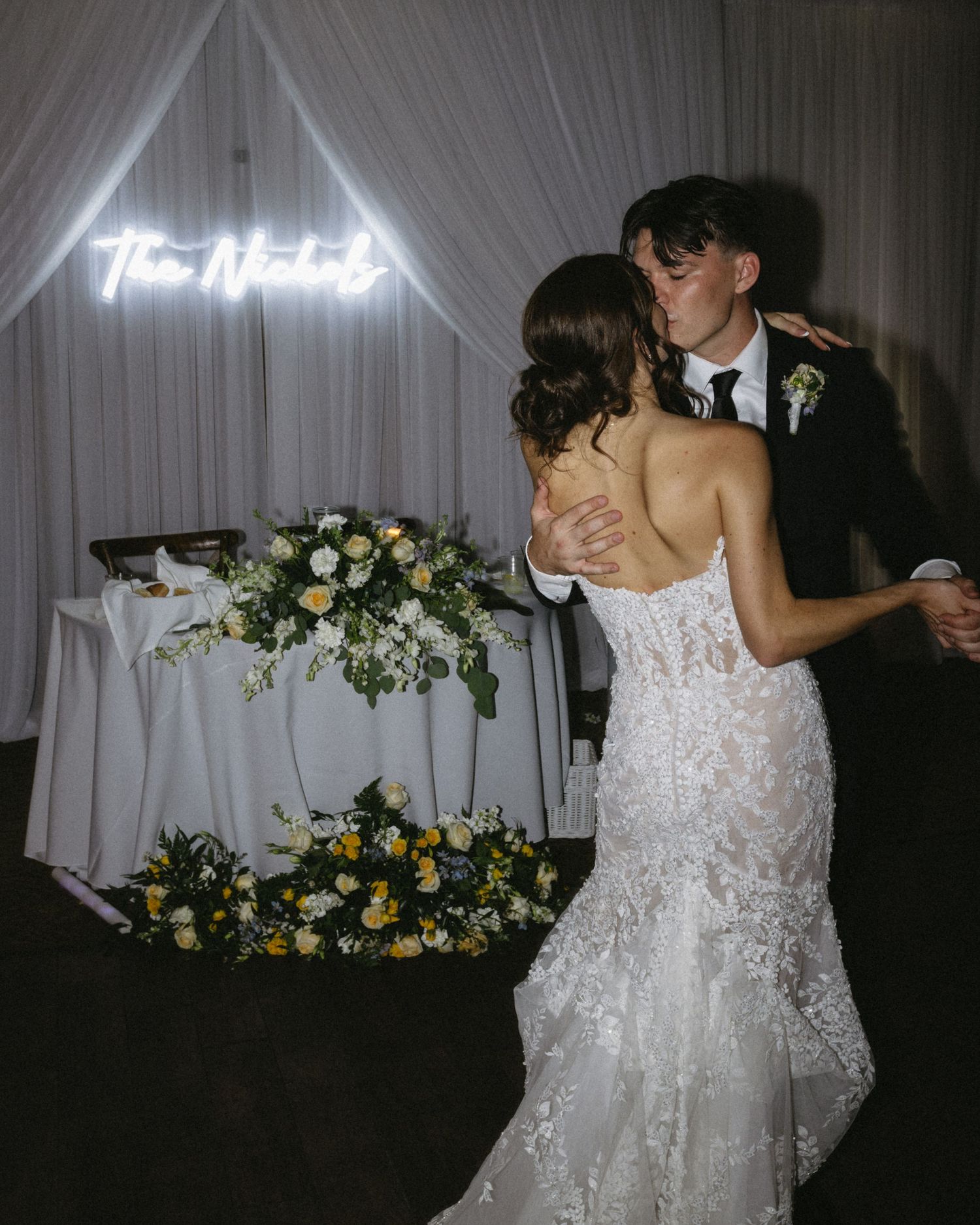 Newlywed couple sharing a first dance kiss in front of a neon sign and floral arrangements at their wedding reception.