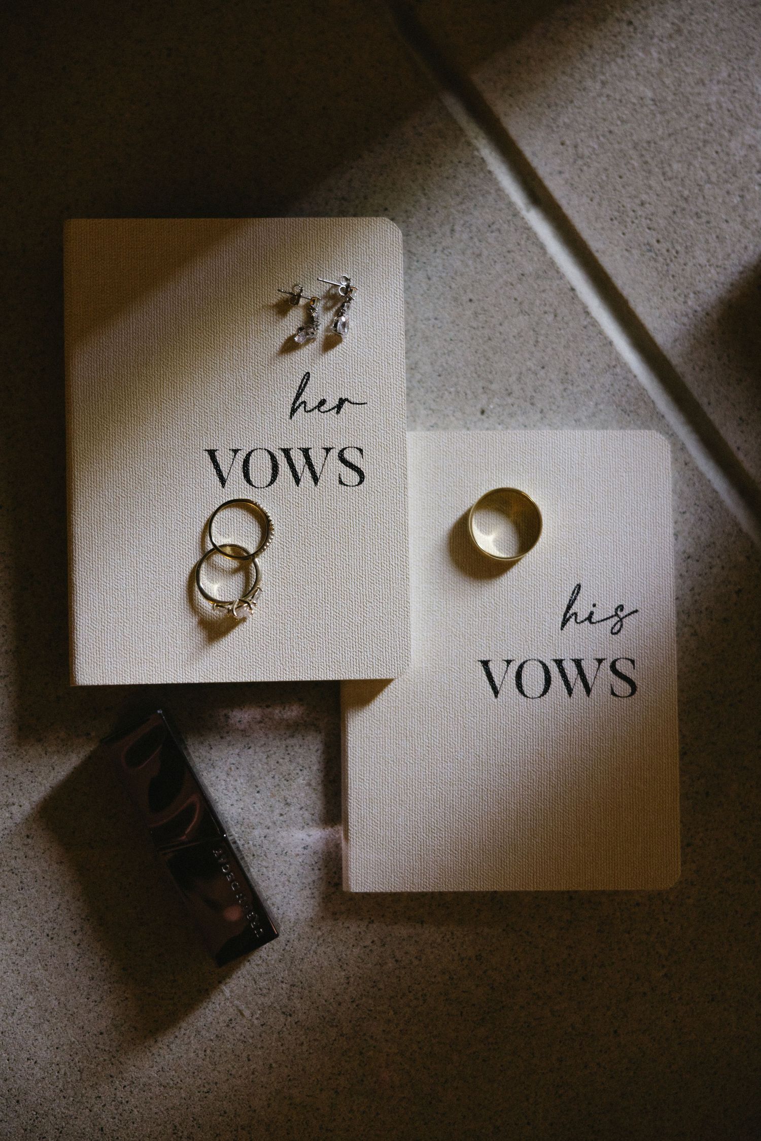 Two elegant vow books labeled his and hers displayed with wedding rings and earrings on a neutral surface.