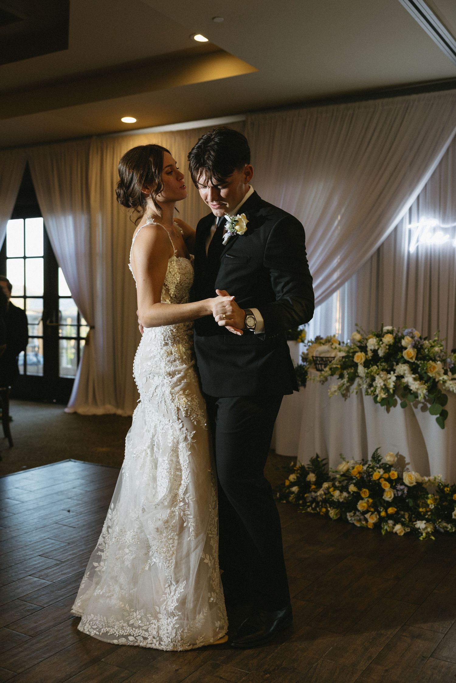 Bride and groom share their first dance in an elegant wedding reception venue adorned with white drapes and yellow floral arrangements.