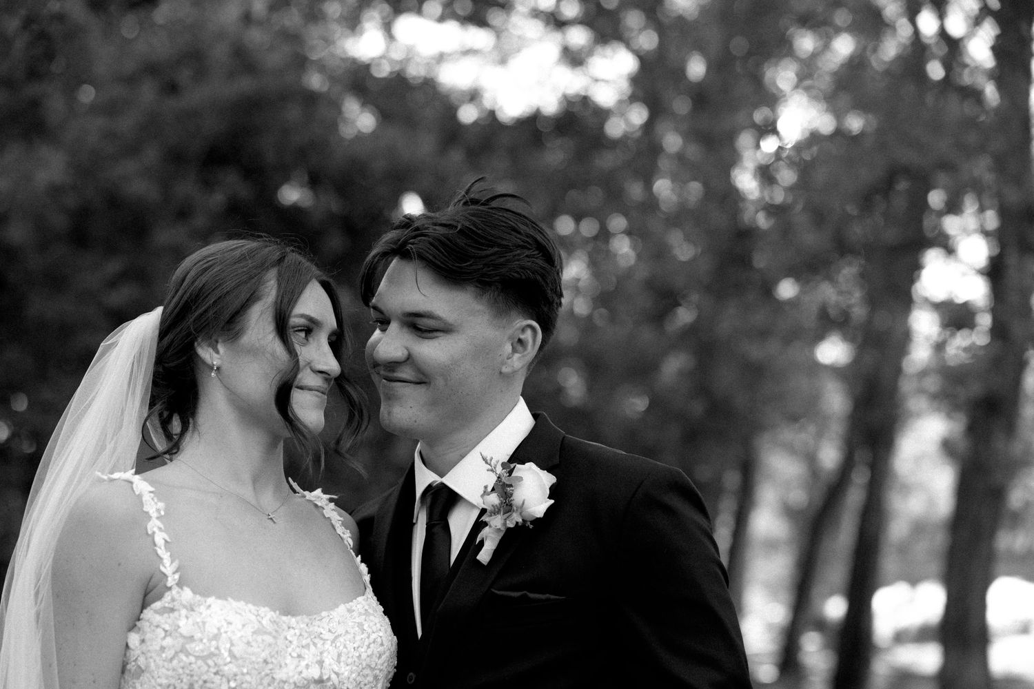 Black and white wedding photo of smiling bride in lace gown and groom in suit with boutonniere outdoors.