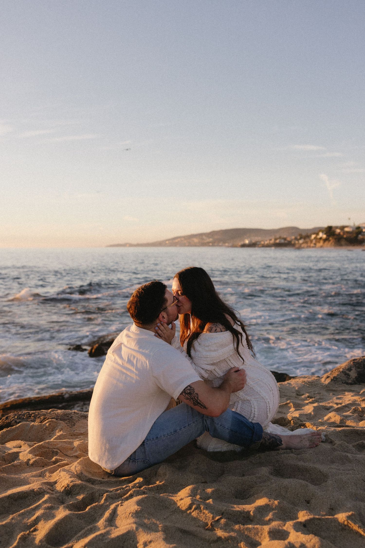 A couple relaxes together on the beach sand at sunset with ocean waves nearby.