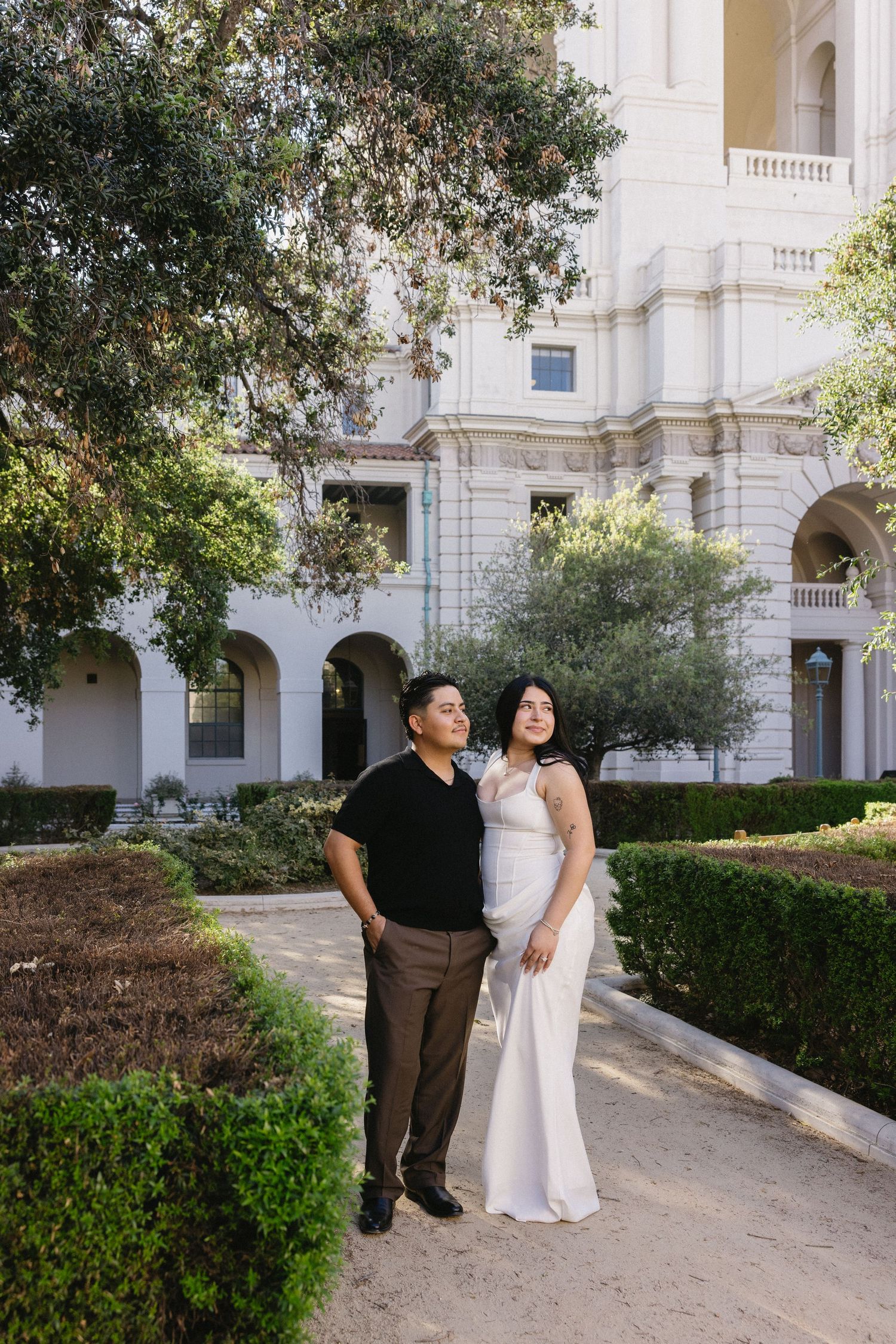 Couple in elegant attire poses in garden of white neoclassical building, woman in white gown, man in black shirt.