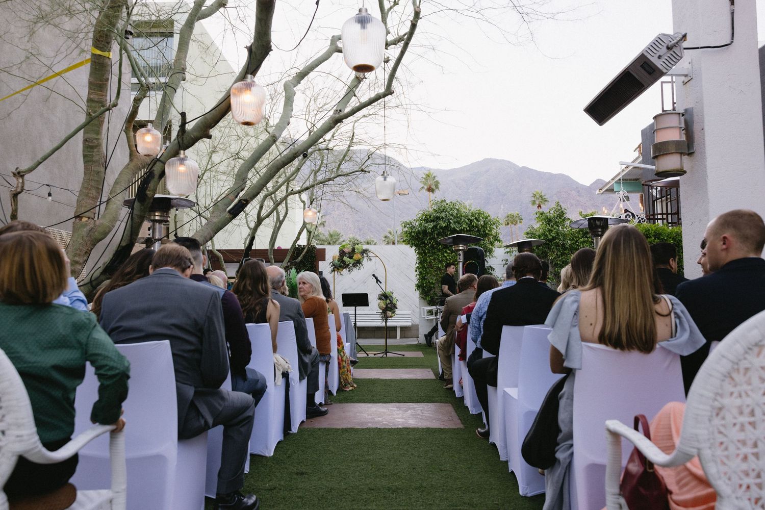 Outdoor wedding ceremony with guests seated in white chairs under trees with mountain views.