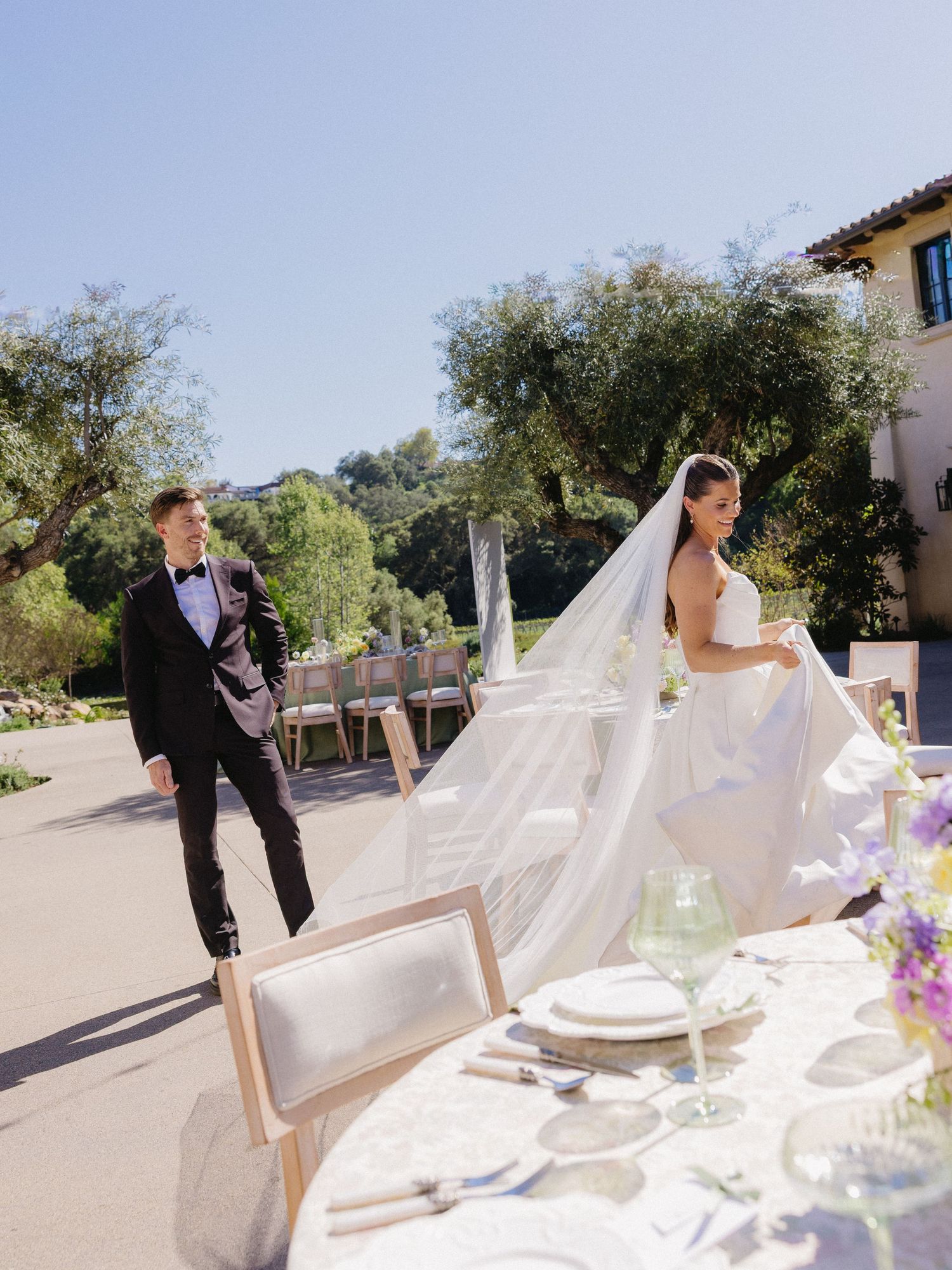 Bride in flowing white gown and veil walks with groom in black tuxedo at elegant outdoor wedding venue.