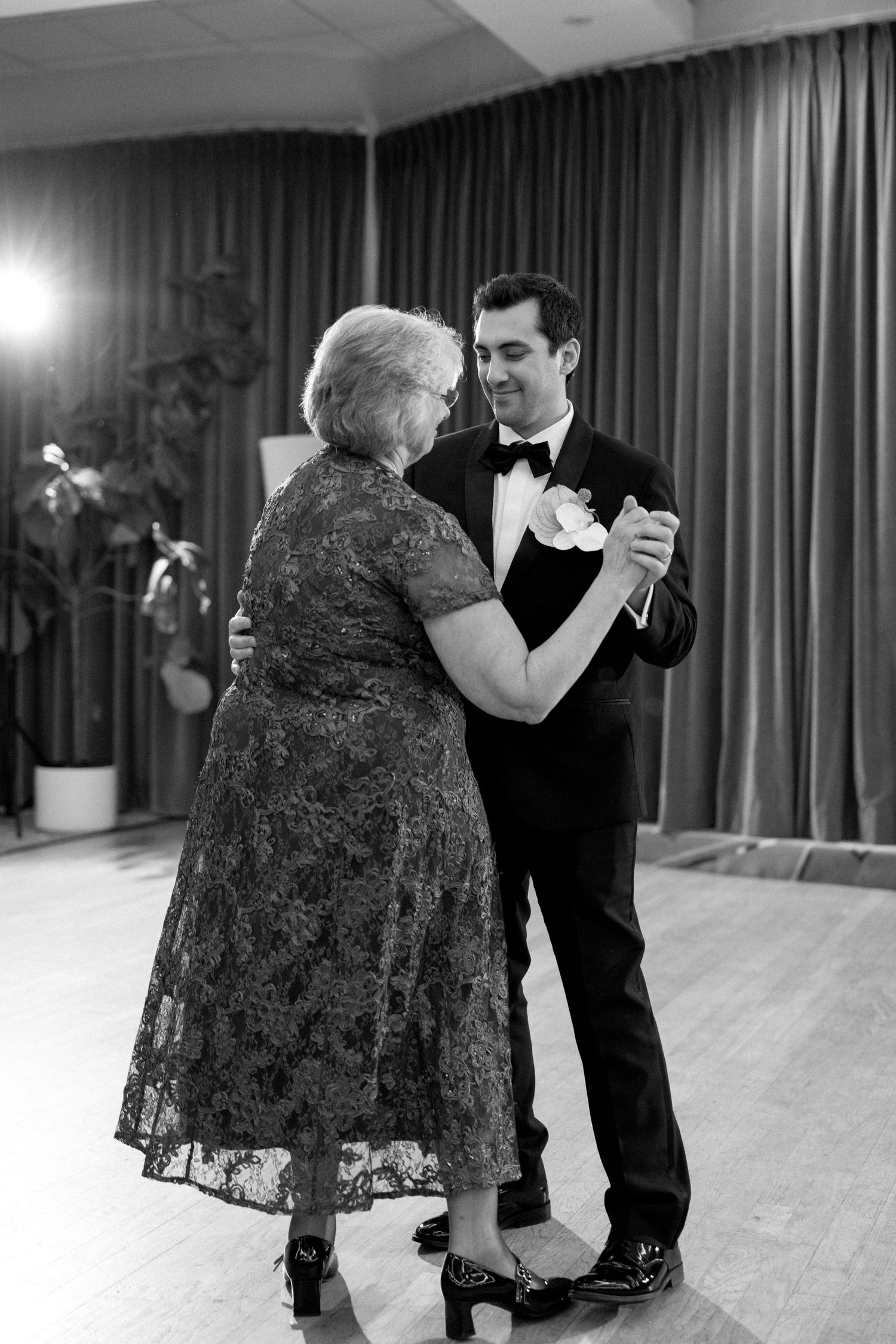 Groom in tuxedo shares mother-son dance with elderly woman in lace dress at wedding reception.