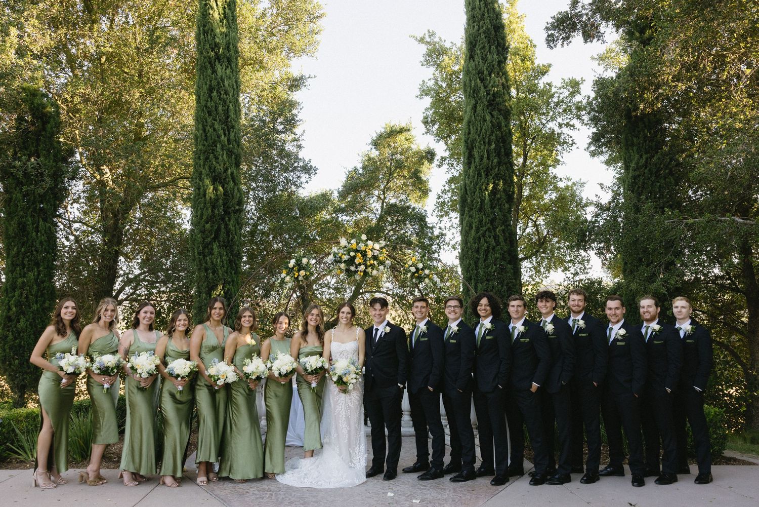 Wedding party posing outdoors with bridesmaids in sage green gowns and groomsmen in black suits among tall cypress trees.