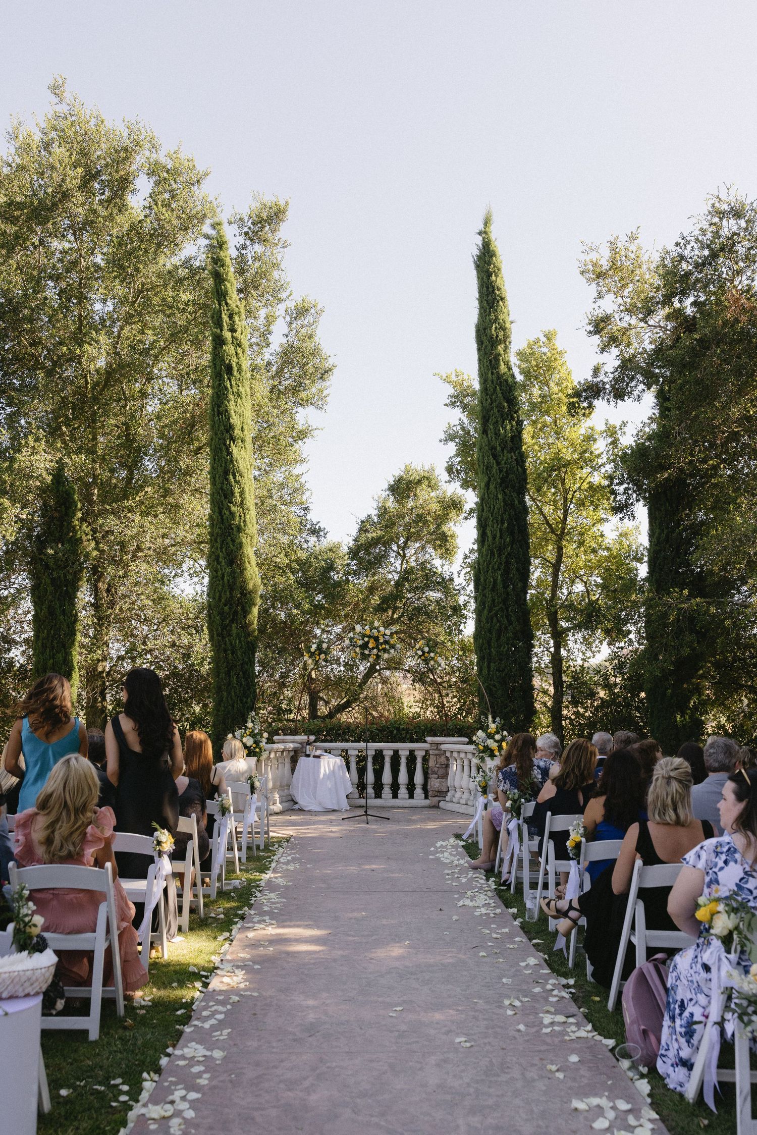 Outdoor wedding ceremony aisle lined with white chairs, cypress trees, and rose petals leading to an elegant garden altar.