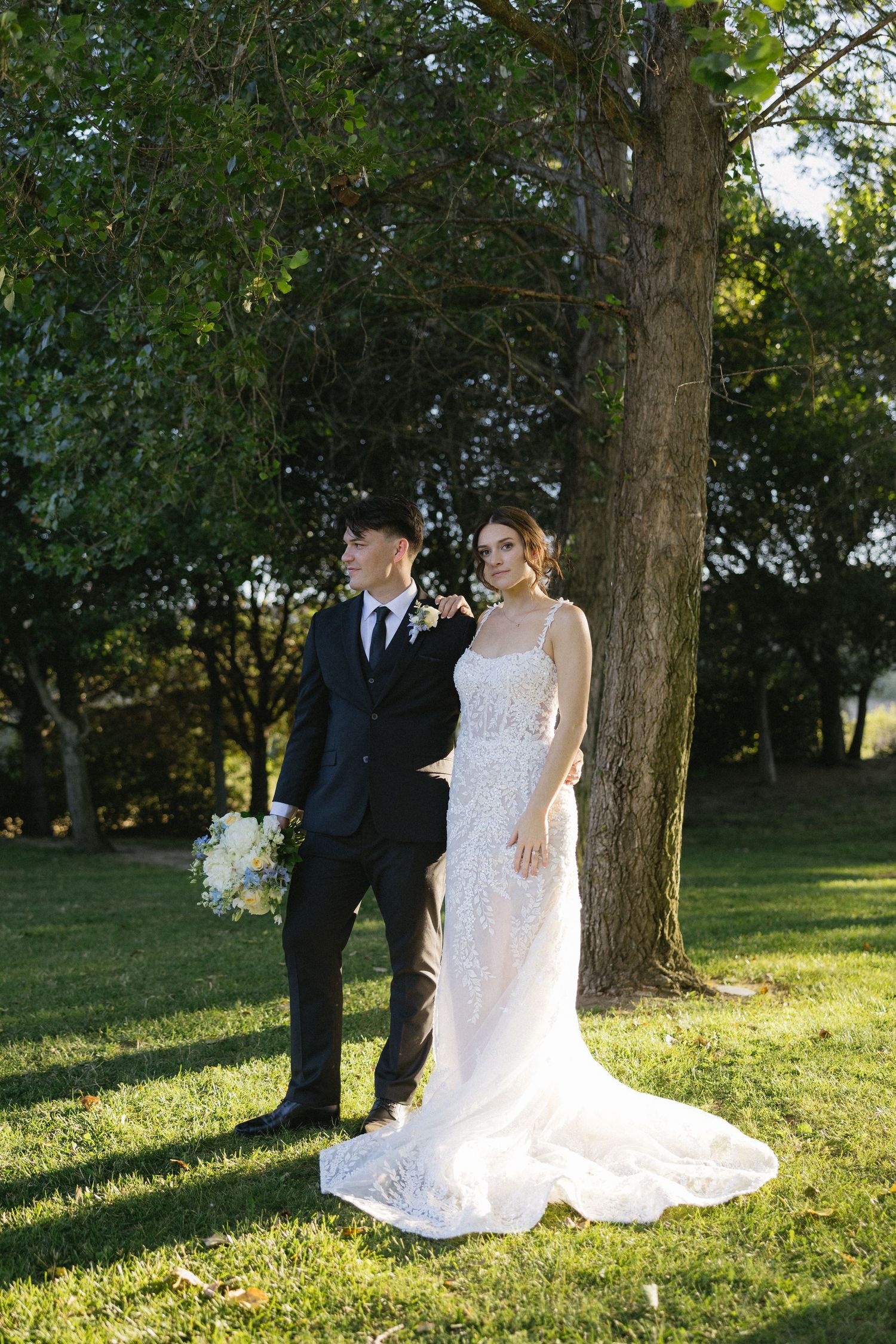 Bride in lace gown and groom in dark suit pose together outdoors near a large tree on green grass.