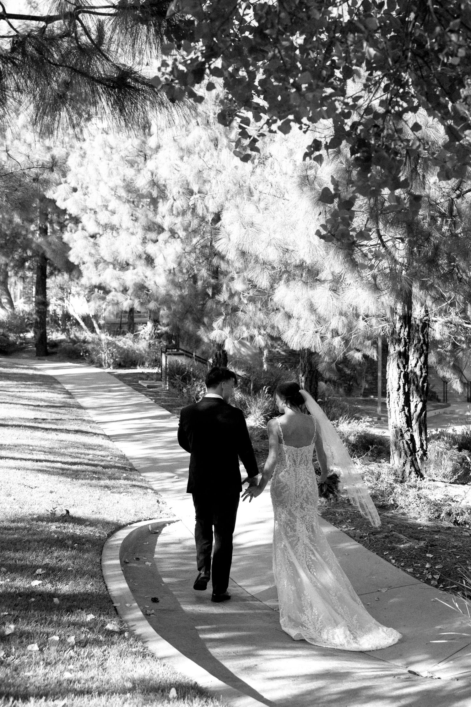 Black and white photo of a bride and groom walking hand in hand down a curved path surrounded by lush trees.