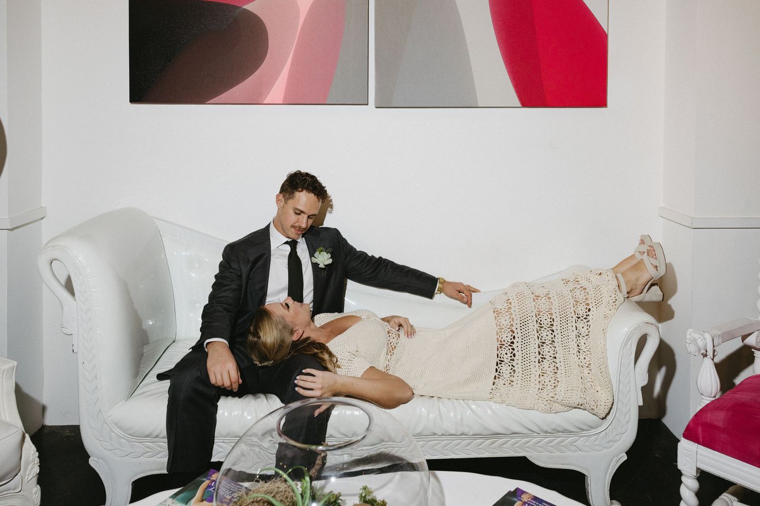 Bride and groom relaxing on a white ornate sofa, with the bride lying across the groom's lap in a wedding dress.