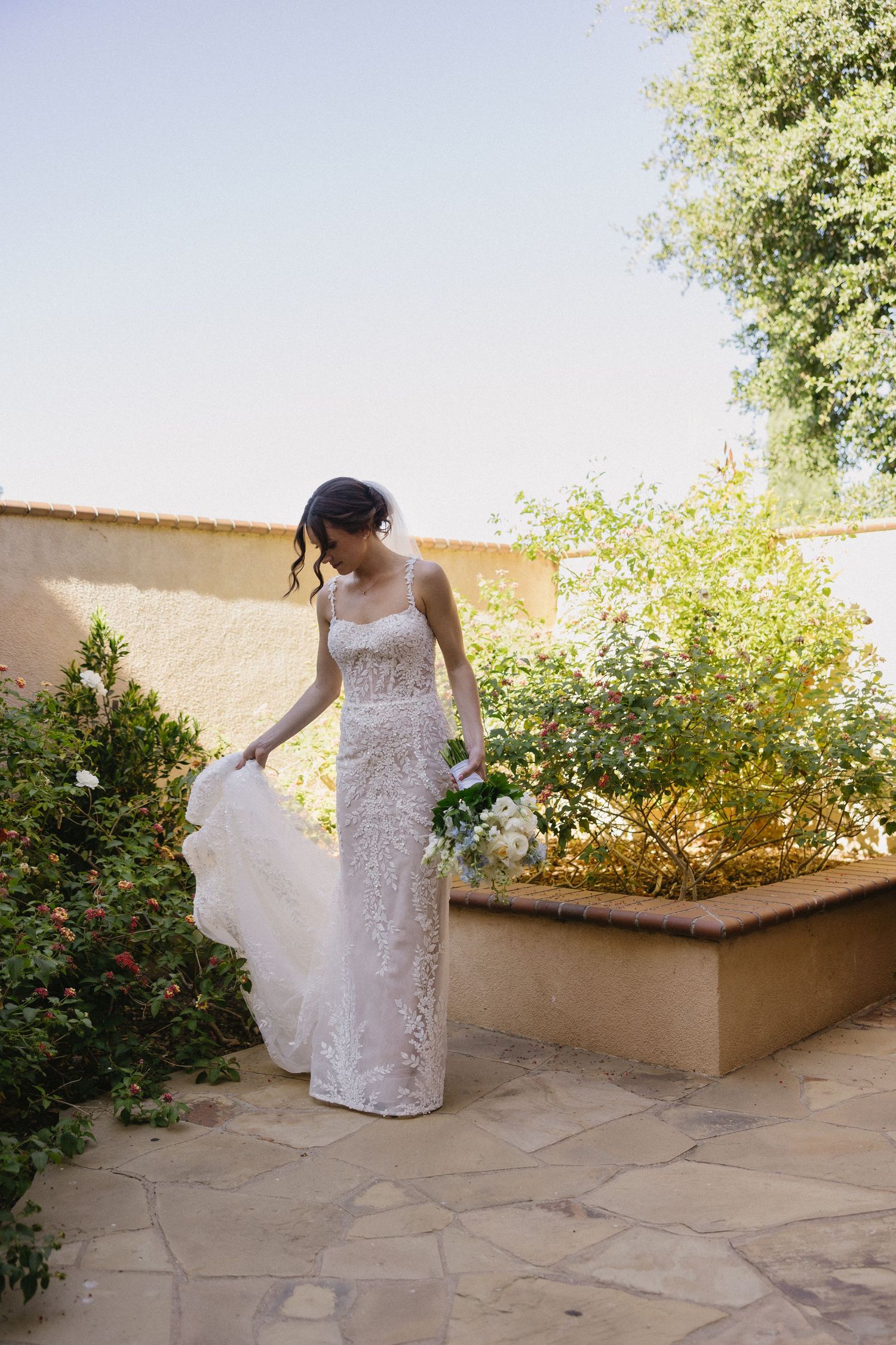 Bride in lace wedding gown holding bouquet, standing in sunlit garden courtyard with green shrubs and stone wall.