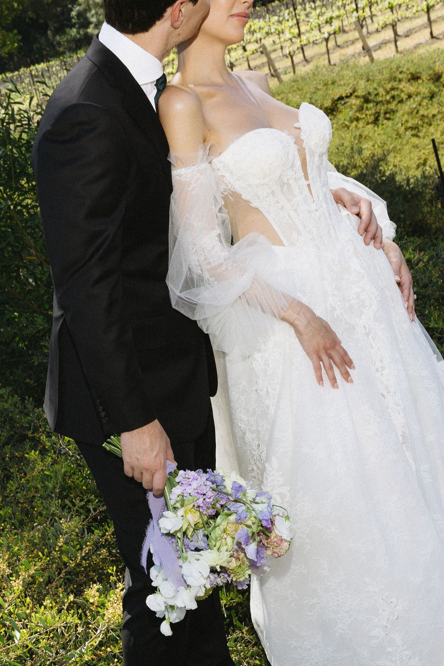 A bride in a white lace gown holds a purple bouquet as a groom in a black tuxedo kisses her cheek outdoors.