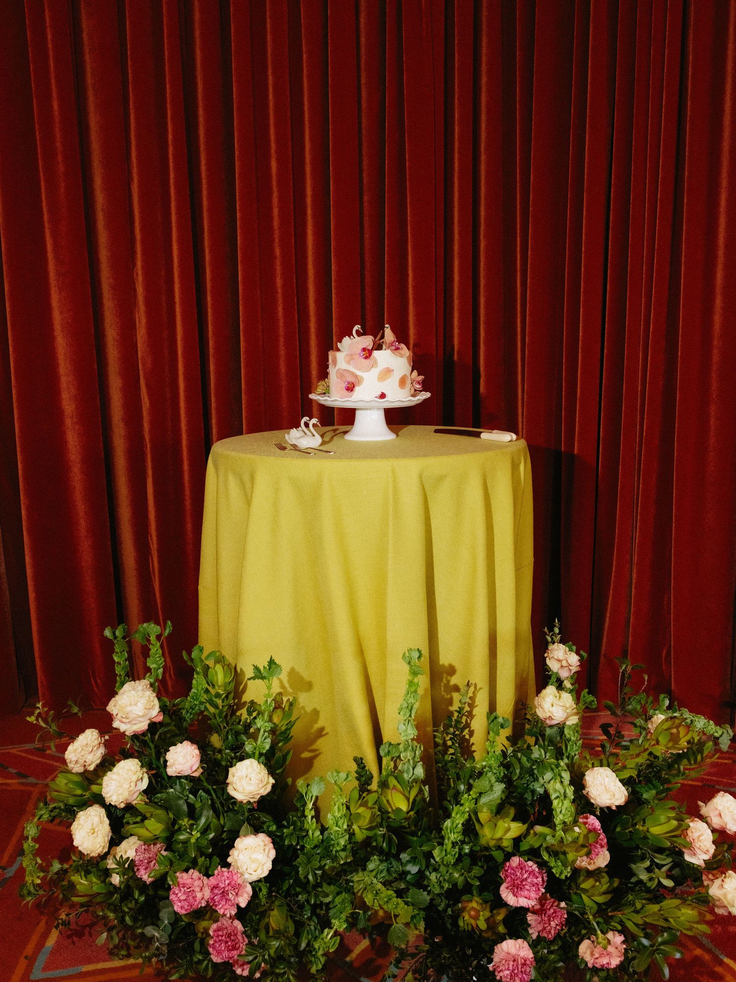 Small wedding cake on white pedestal atop yellow draped table, surrounded by pink and white floral arrangements with red curtain backdrop.