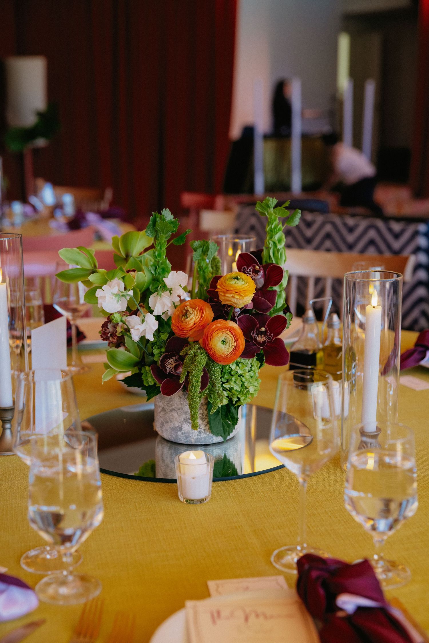 Elegant wedding reception table with yellow linen, floral centerpiece featuring orange ranunculus, burgundy orchids, and green foliage.