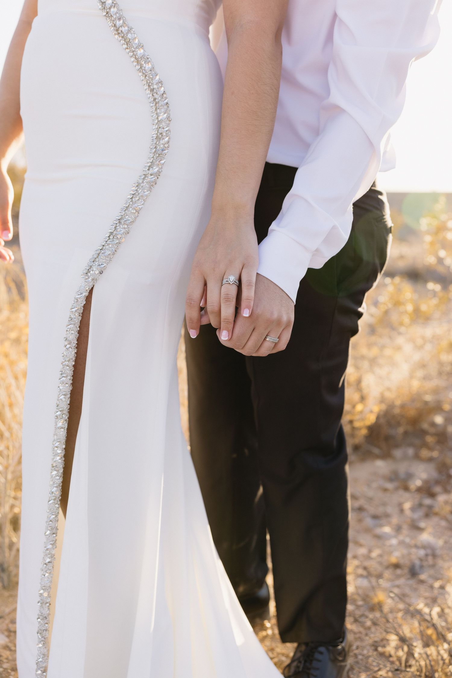 Close up of a white wedding dress with crystal embellishments as couple holds hands in desert setting.