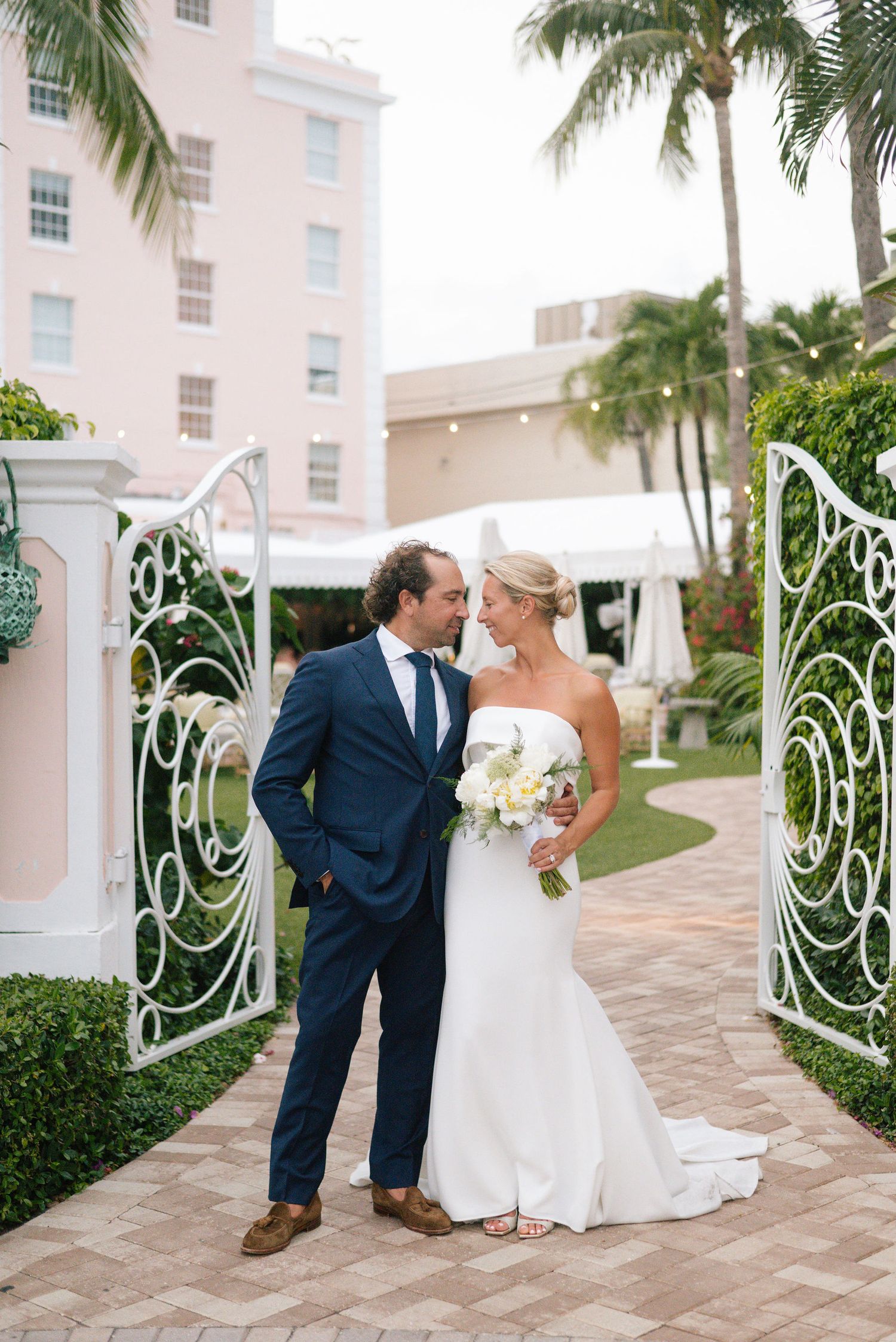 Wedding couple sharing playful moments on brick pathway near pink building with palm trees.