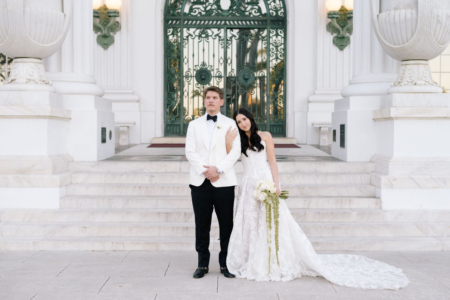 A wedding couple poses on white steps with decorative railings and columns.