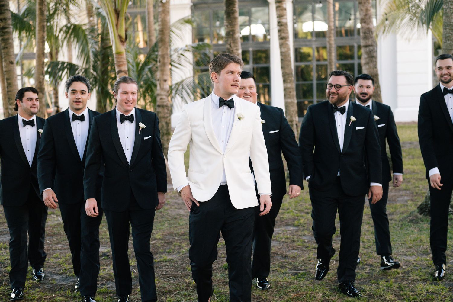 Groomsmen in black tuxedos and one in white stand together at an outdoor wedding venue.