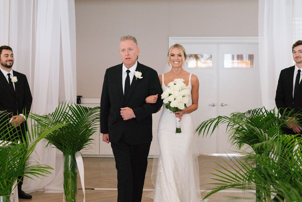 Wedding ceremony in a bright white venue with tropical palm decor and elegant floral arrangements.