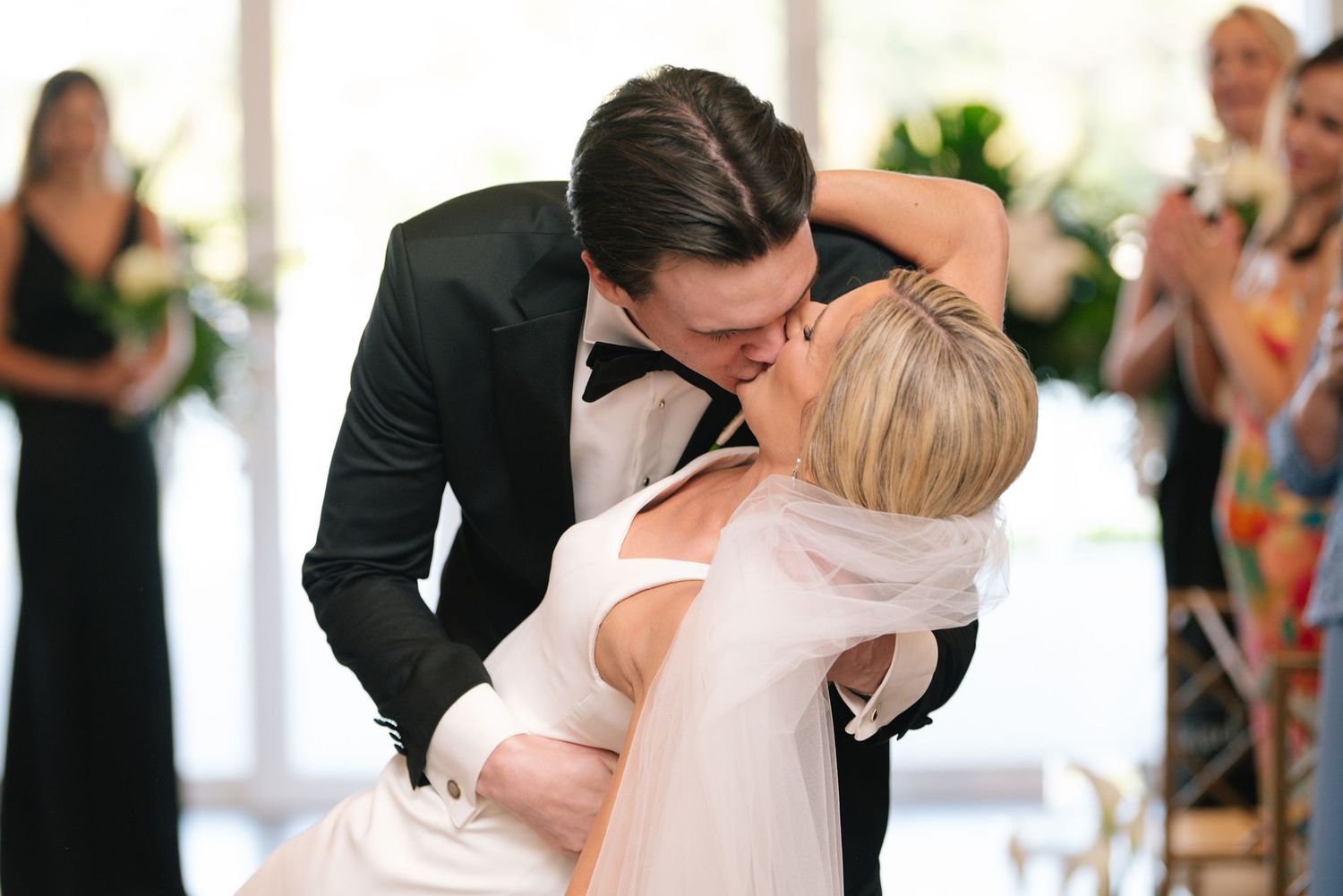 Romantic first kiss moment during wedding ceremony with guests looking on.