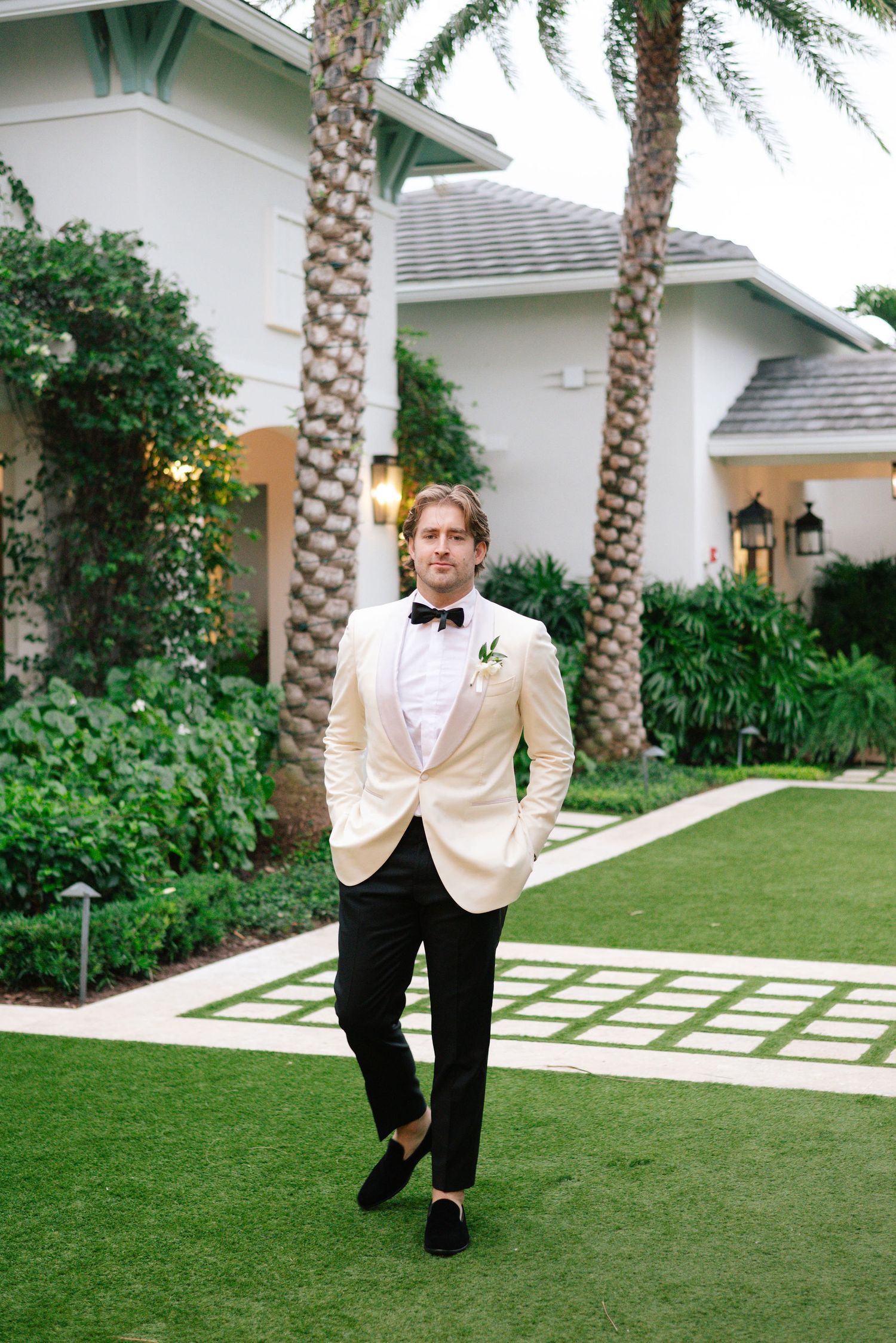 Groom in cream tuxedo poses playfully on lawn with palm trees.