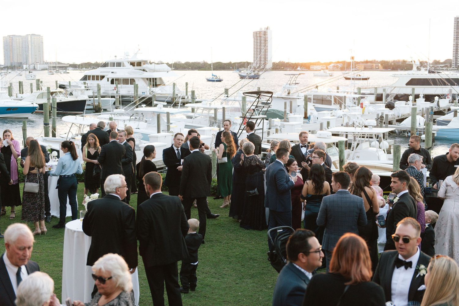 Elegant wedding reception crowd gathered on a waterfront lawn with marina and sunset views.