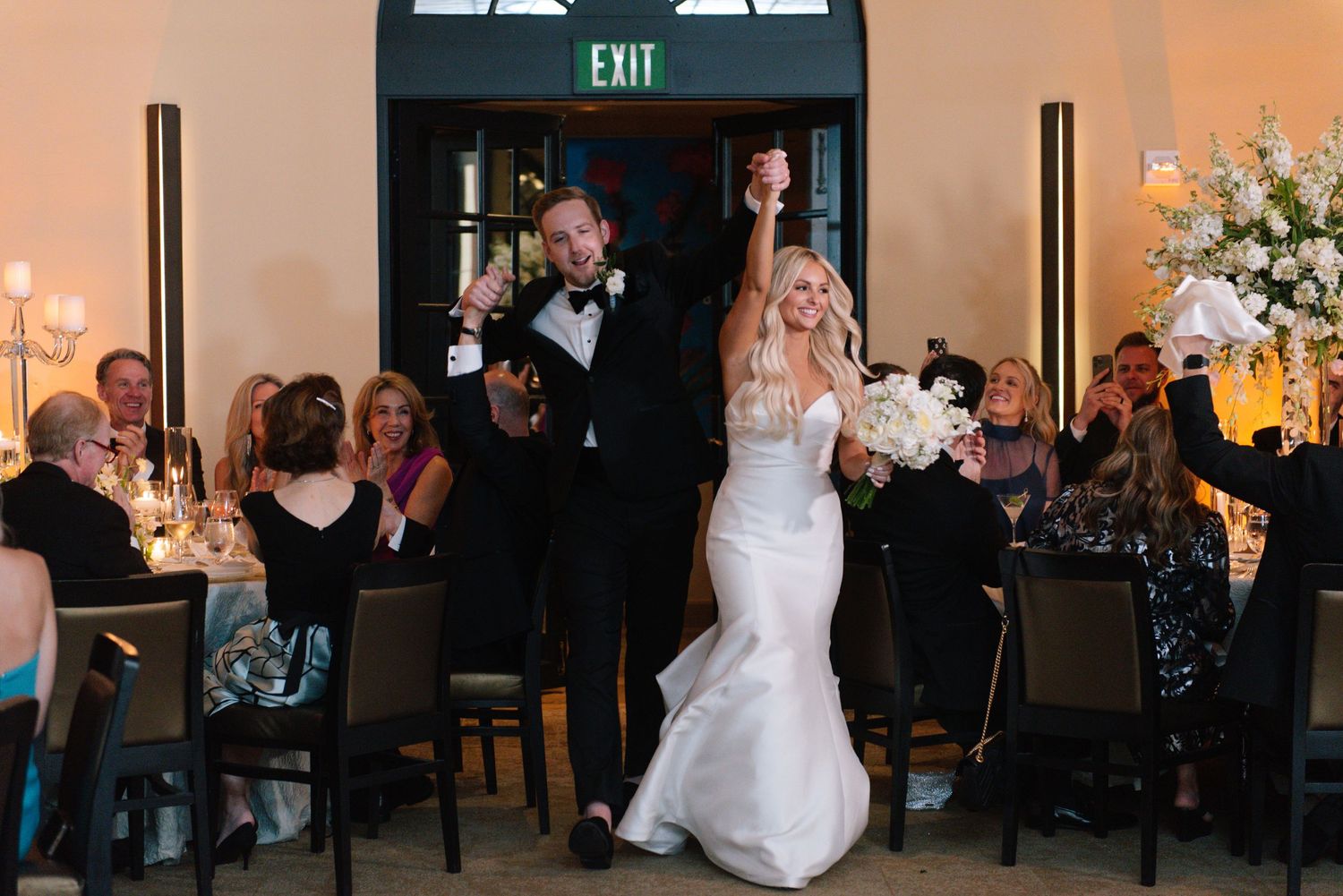 A couple makes their grand entrance at a wedding reception as guests seated at tables cheer and applaud.
