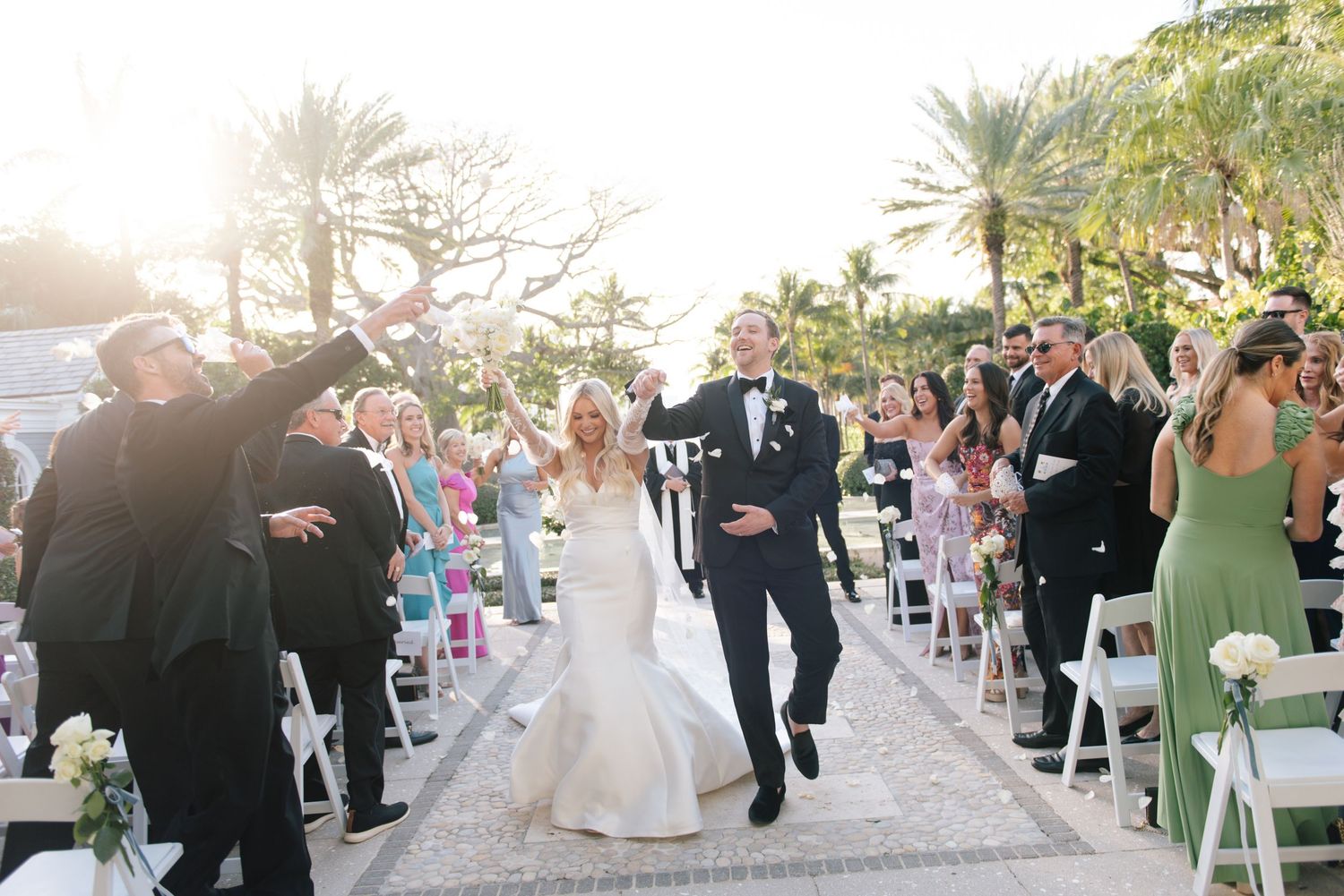 Newlyweds joyfully exit their ceremony while guests celebrate along palm-lined aisle with white chairs.