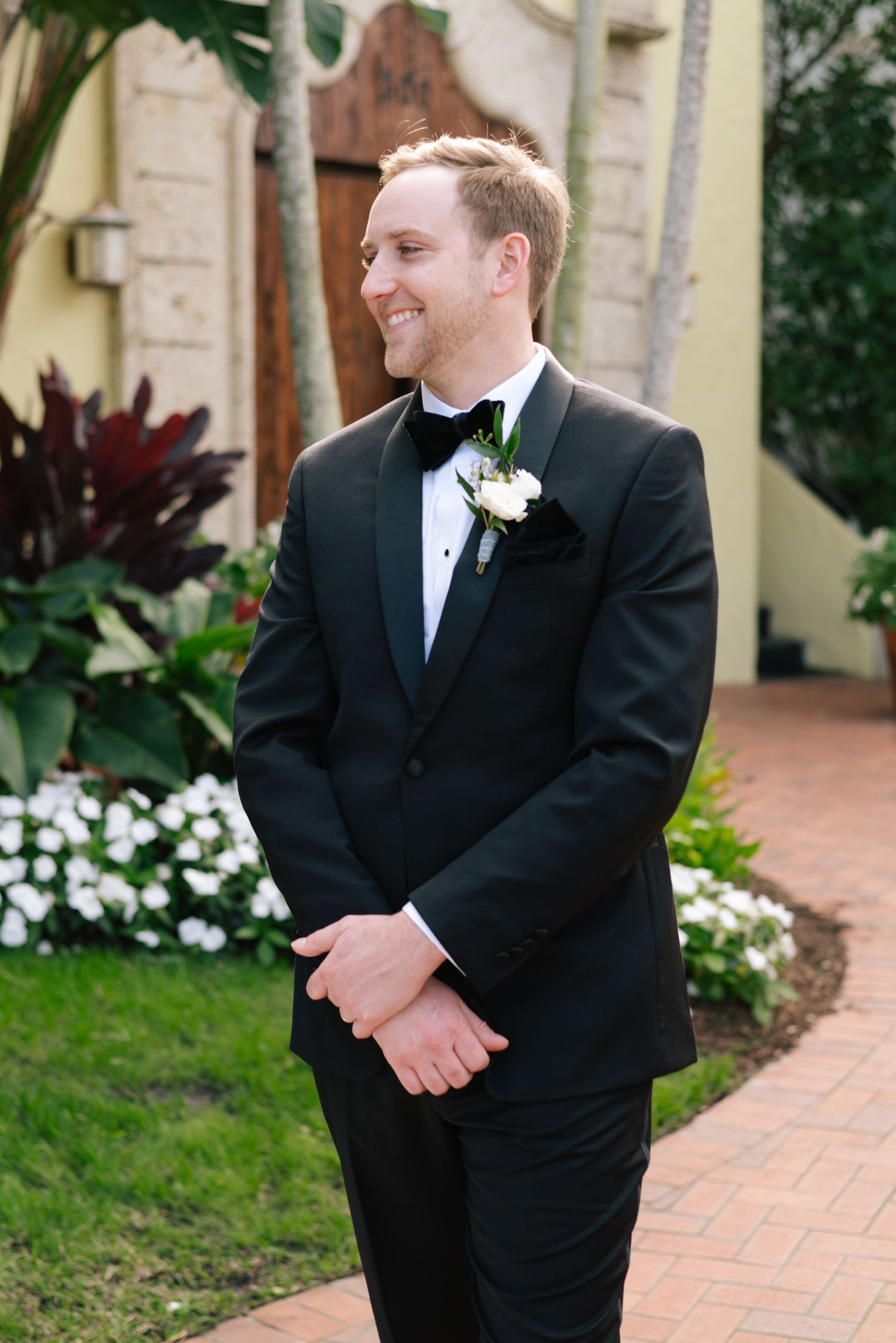 Wedding party members in black tuxedos stand along brick pathway surrounded by landscaping.