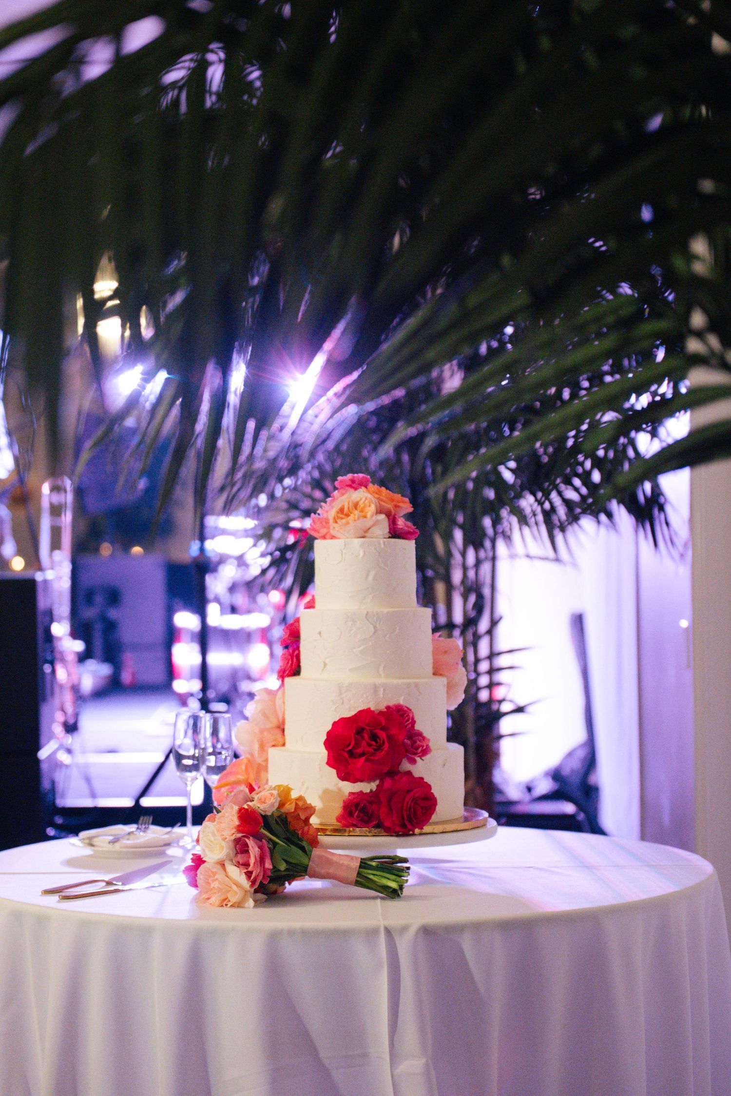 White tiered wedding cakes adorned with pink and red flowers displayed on tables with dramatic lighting.