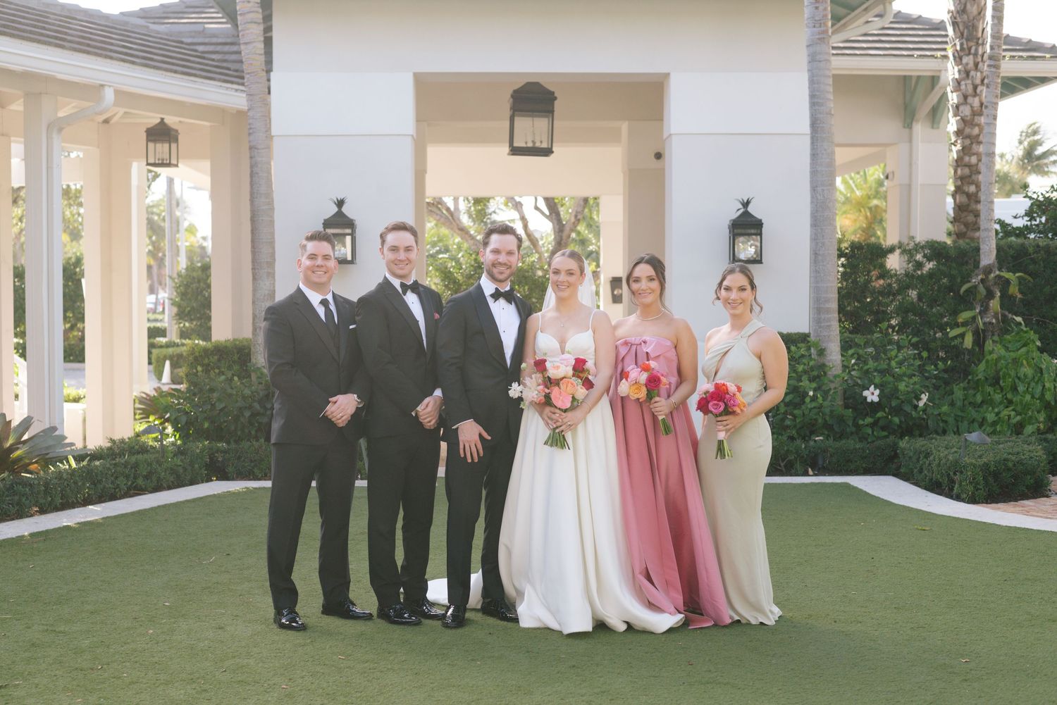 Wedding party members in pink dresses and dark suits pose together on a manicured lawn.