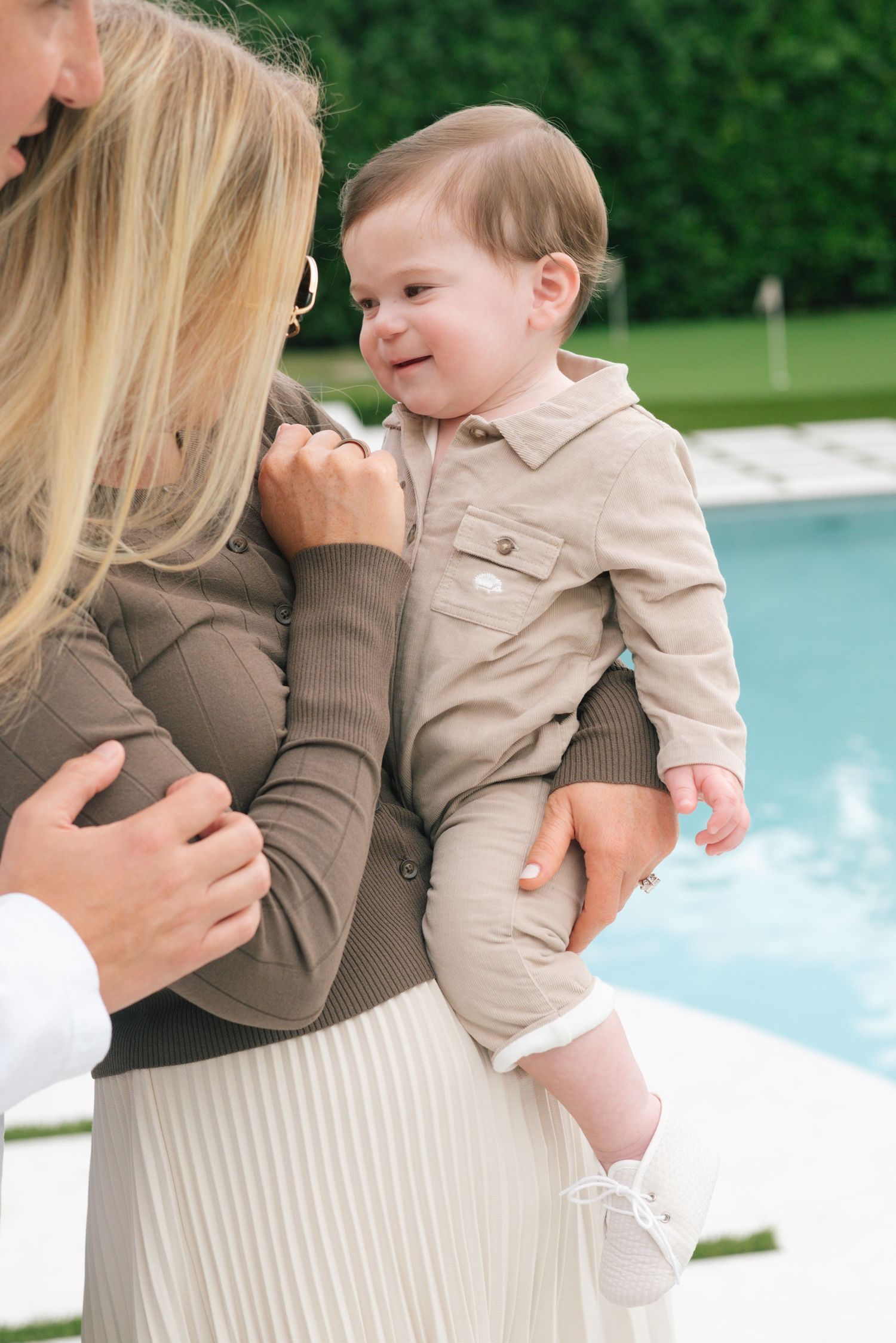 A toddler in beige outfit smiles while being held near a swimming pool on a sunny day.