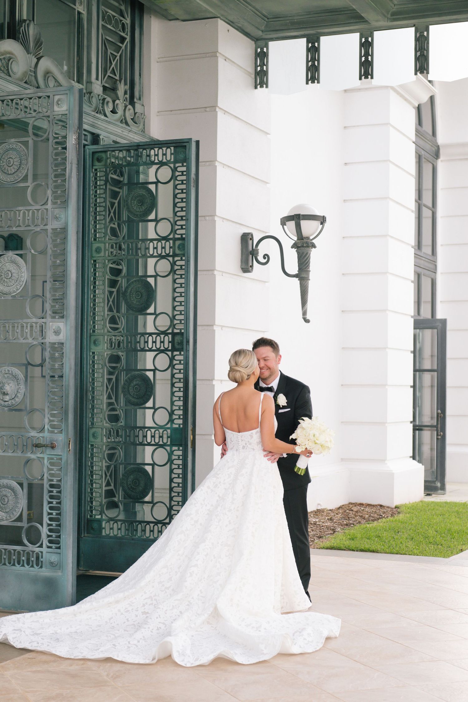 Bride and groom share a romantic moment outside elegant white venue with ornate green doors.