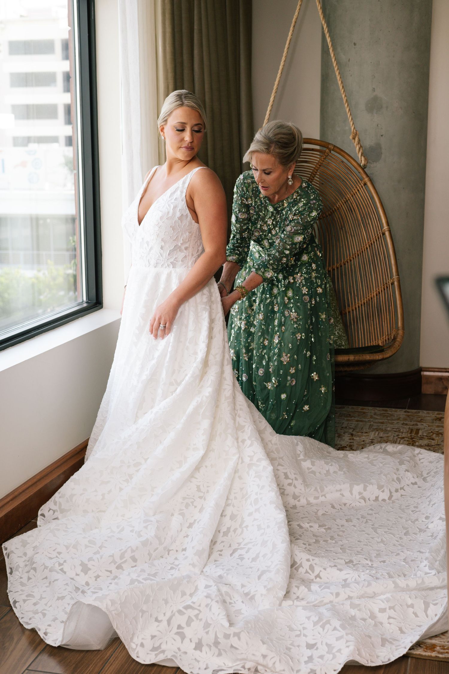 Bride in white lace gown being helped by woman in green floral dress near window.