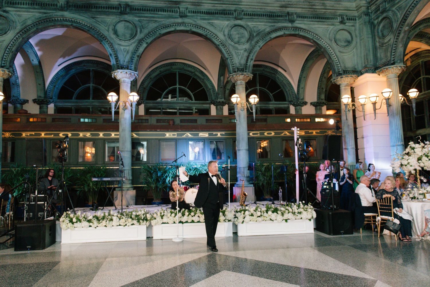 Groom standing in grand ornate ballroom during elegant black tie wedding reception.