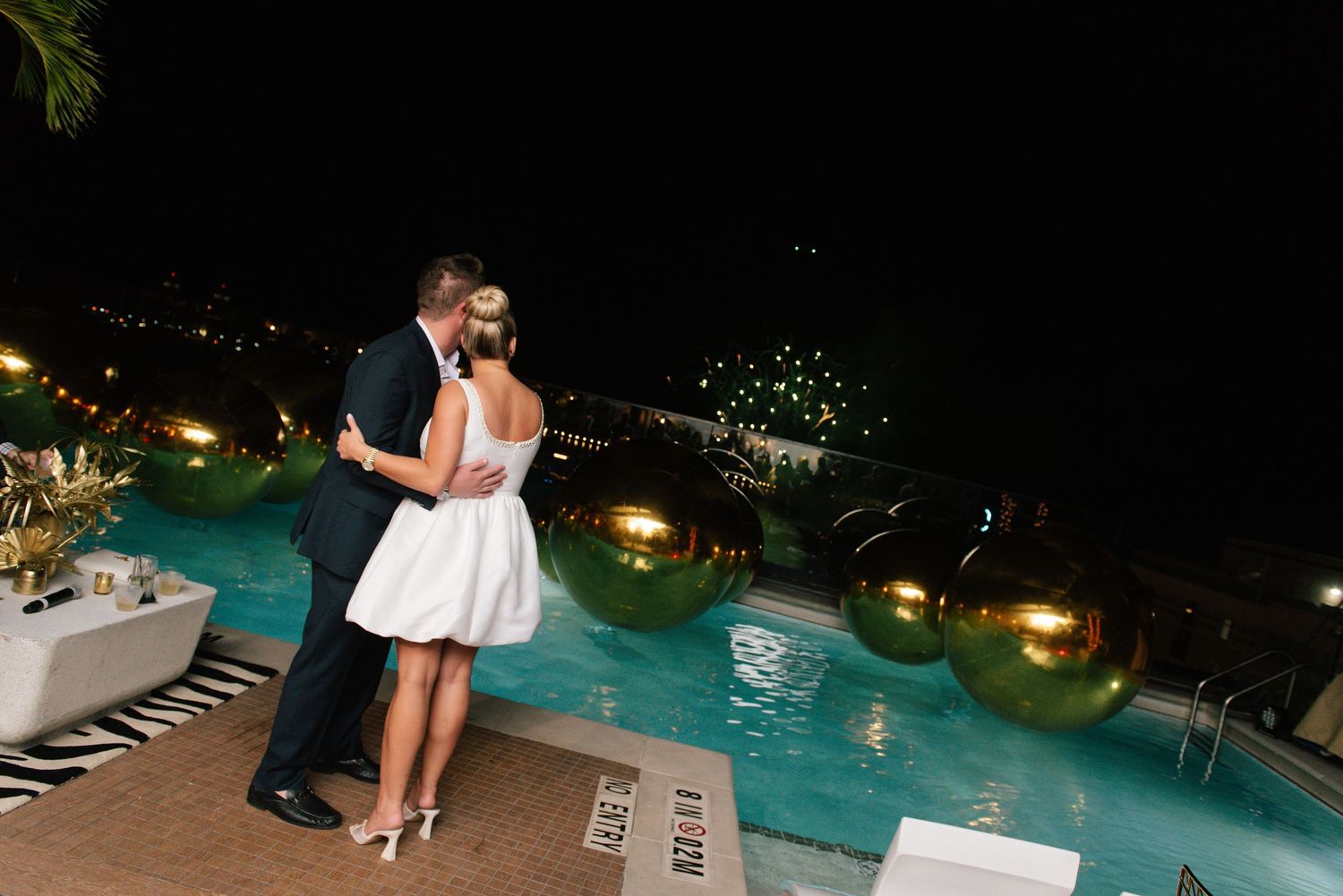 A romantic moment beside an illuminated pool with city lights twinkling in the background.