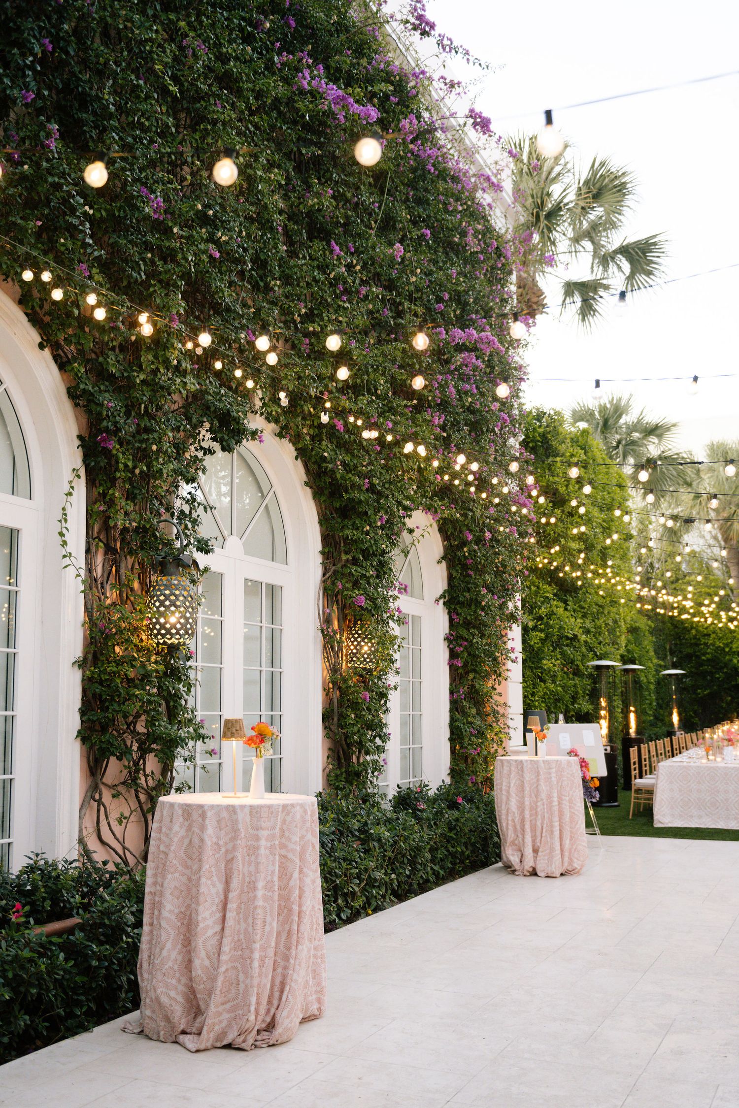 Outdoor wedding venue with string lights, pink draping, and climbing purple flowers.